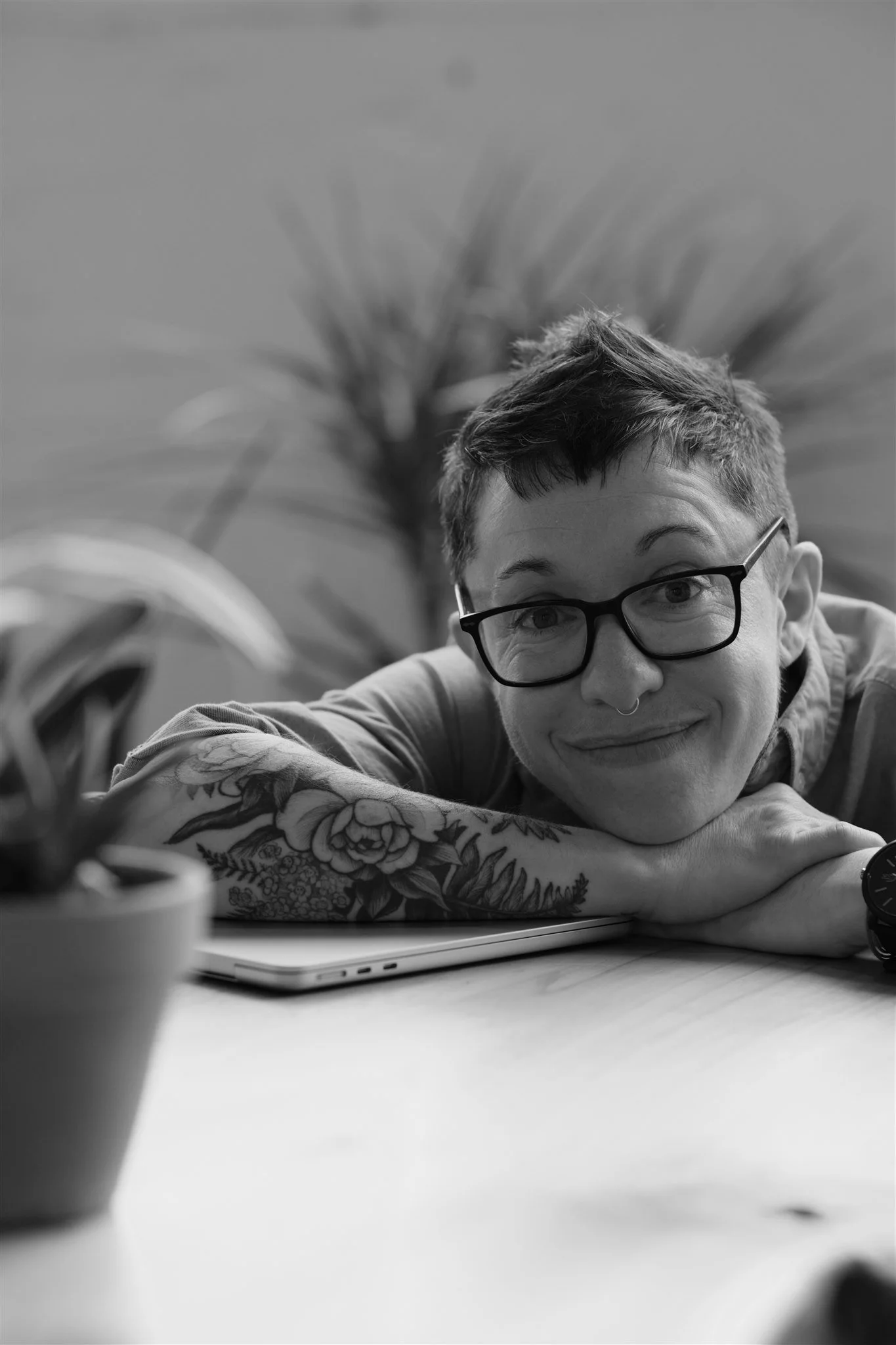 Black and white photo of a person with short hair, glasses, and tattoos, resting their head and arms on a table next to a laptop, smiling at the camera, with a potted plant in the foreground.