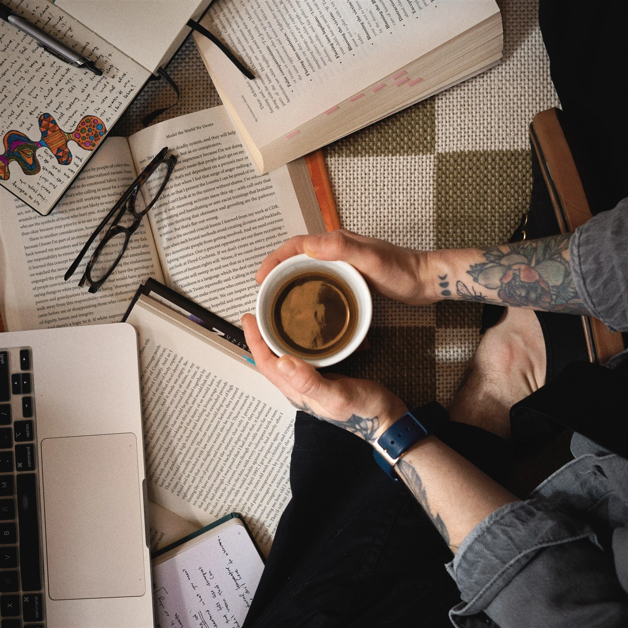 A person holding a cup of coffee over an open book on a table with other books, glasses, a notebook, and a laptop nearby.