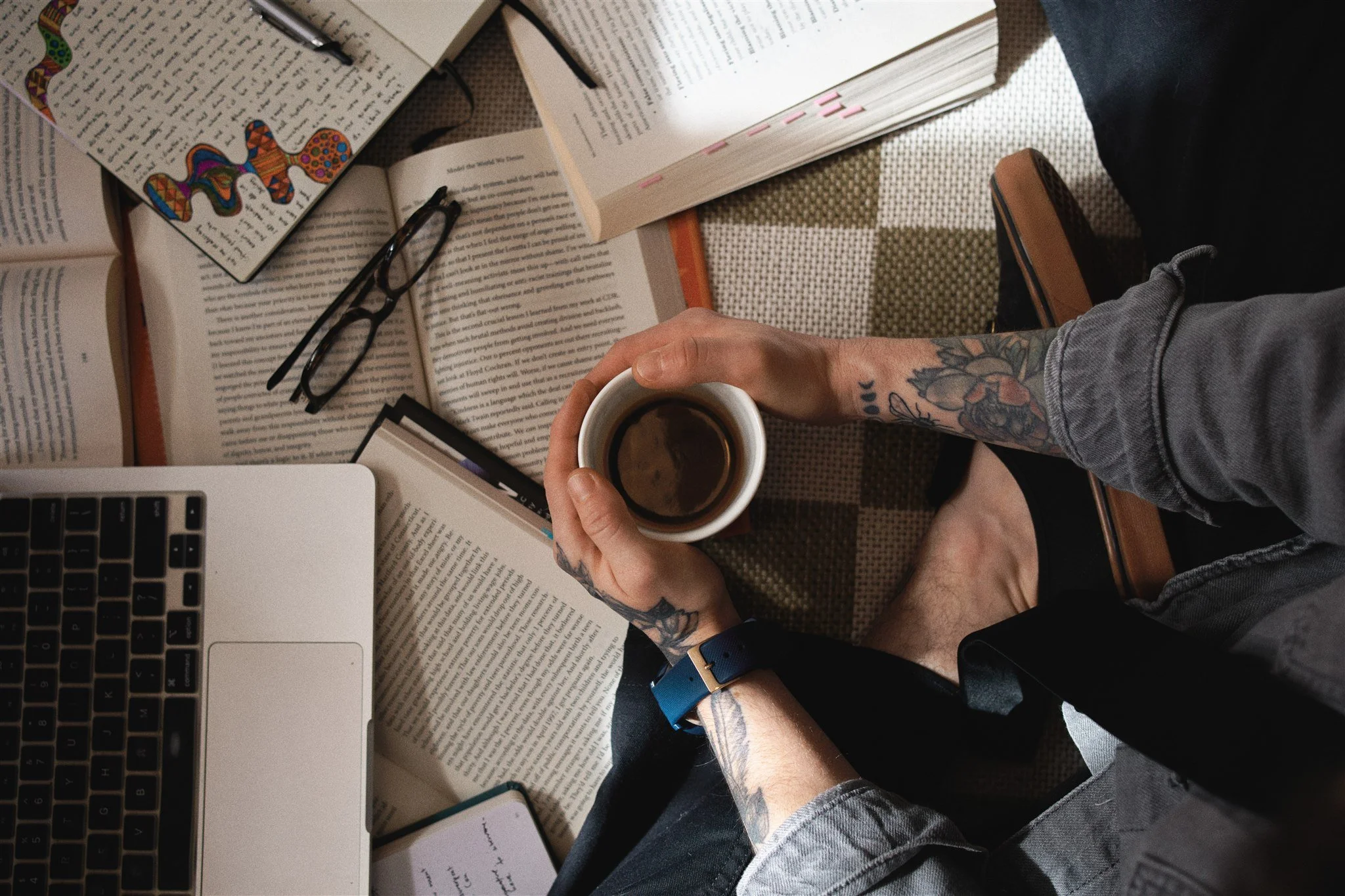 Person holding a cup of coffee or tea among open books, reading glasses, a laptop, and a smartphone on a checkered surface.