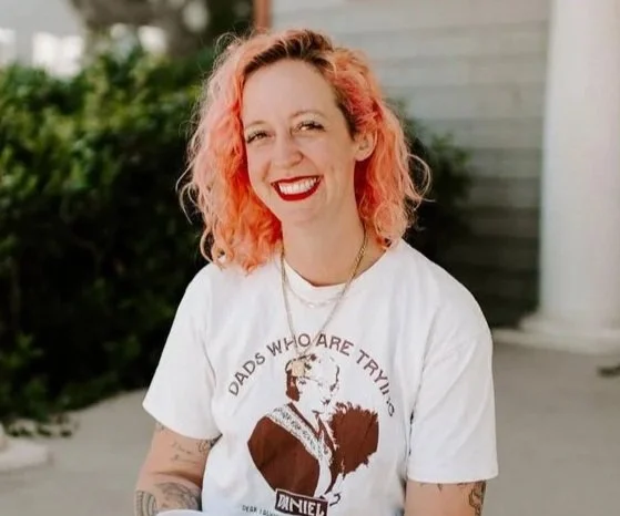 A smiling woman with curly, pinkish-orange hair, wearing a white T-shirt with a printed design, standing outdoors with a blurred background of greenery and a building.