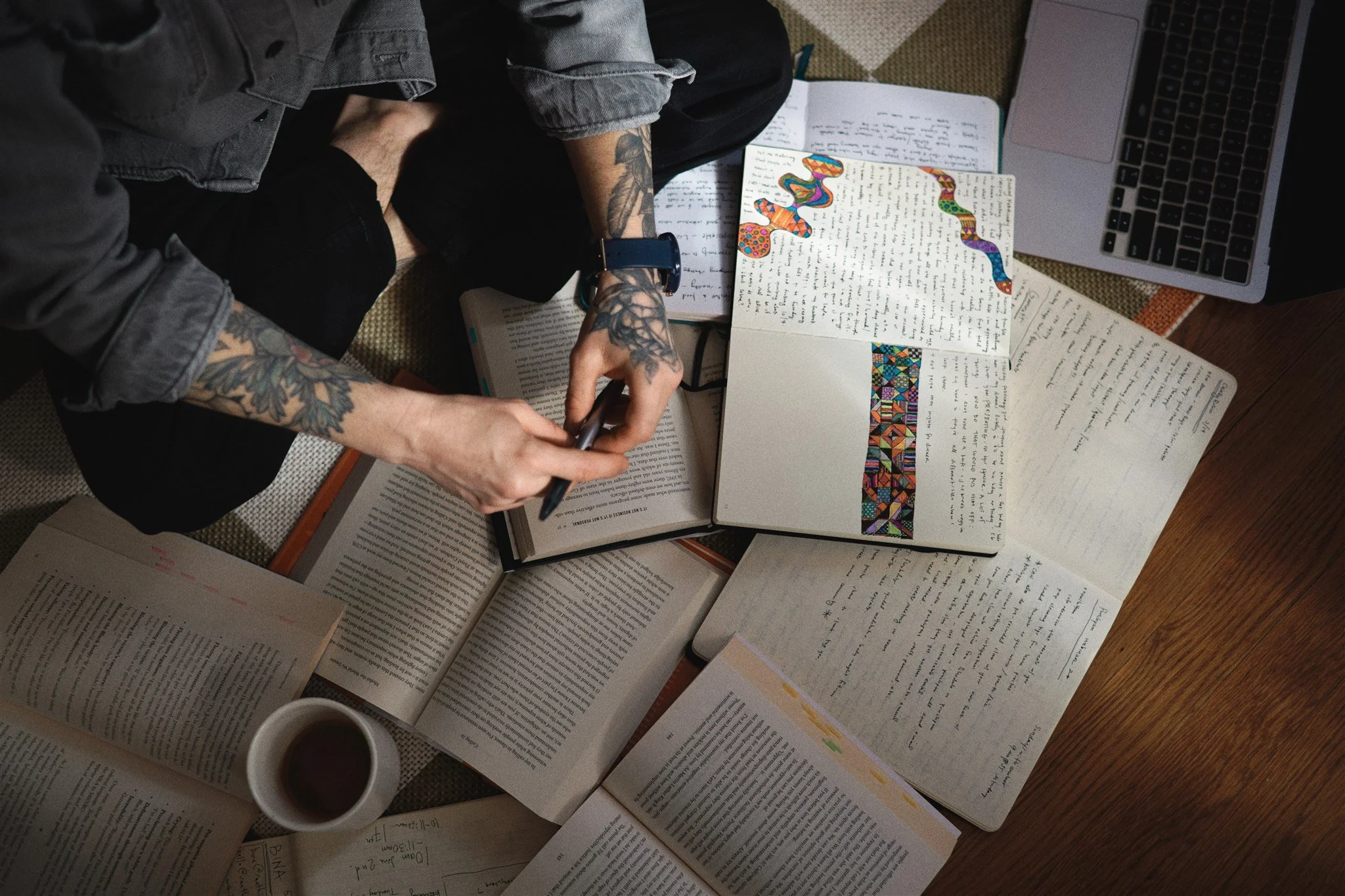 Top-down view of a person with tattooed arms, sitting on the floor, writing in a notebook surrounded by open books, some with colorful doodles or highlighted text, a laptop, and a cup of coffee.