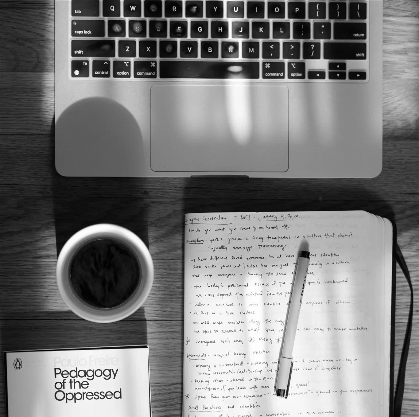 Top-center: A laptop keyboard and trackpad on a wooden desk. Bottom-center: An open notebook with handwritten notes, a pen resting on it, and a cup of coffee. Bottom-left: A book titled "Pedagogy of the Oppressed" by Paulo Freire.