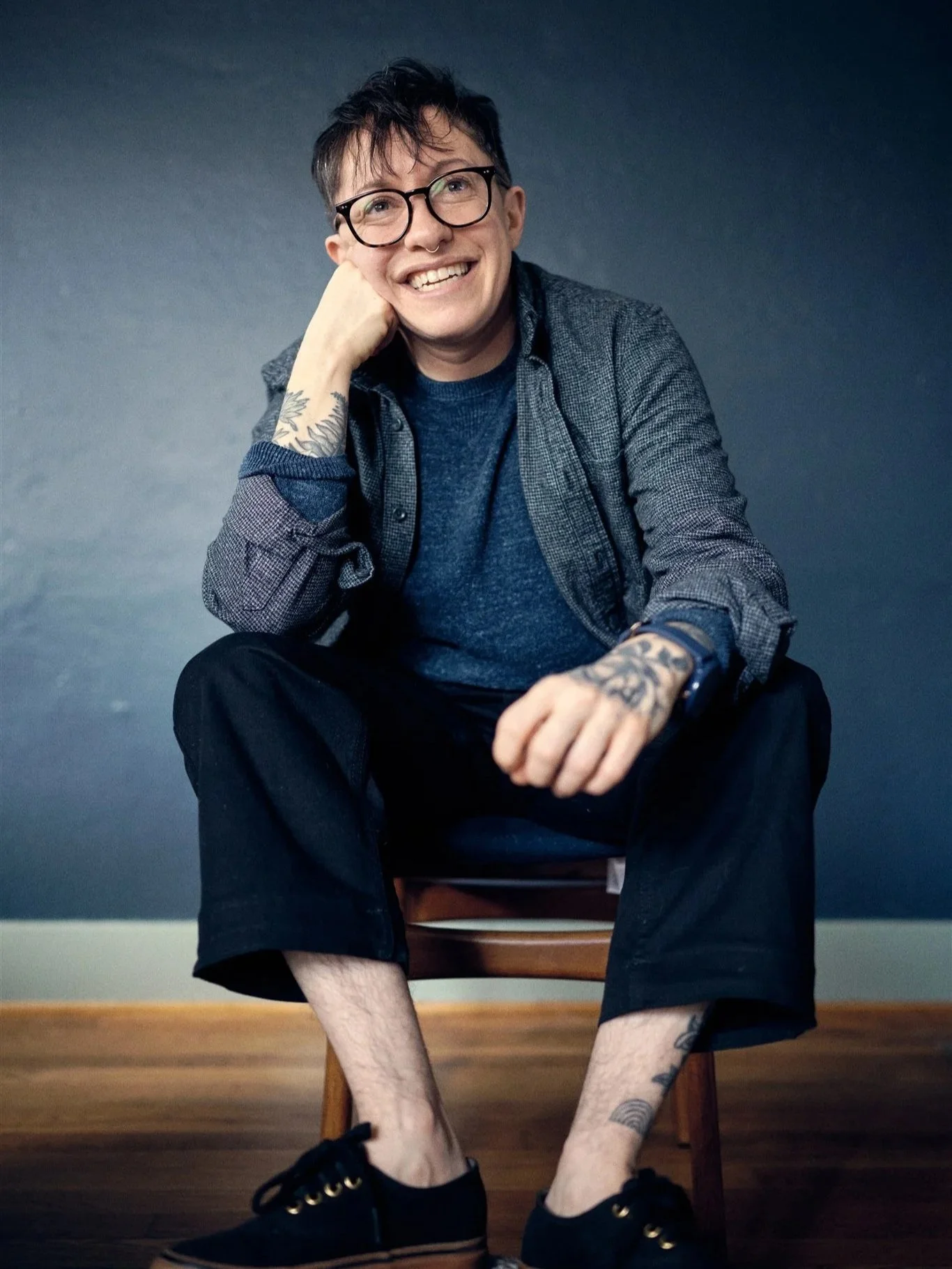 Young person with short dark hair, glasses, and tattoos sitting on a wooden chair against a dark blue wall. He is smiling, resting his head on his left hand, wearing a dark shirt and jeans, with sneakers, and has tattoos on his arms and legs.