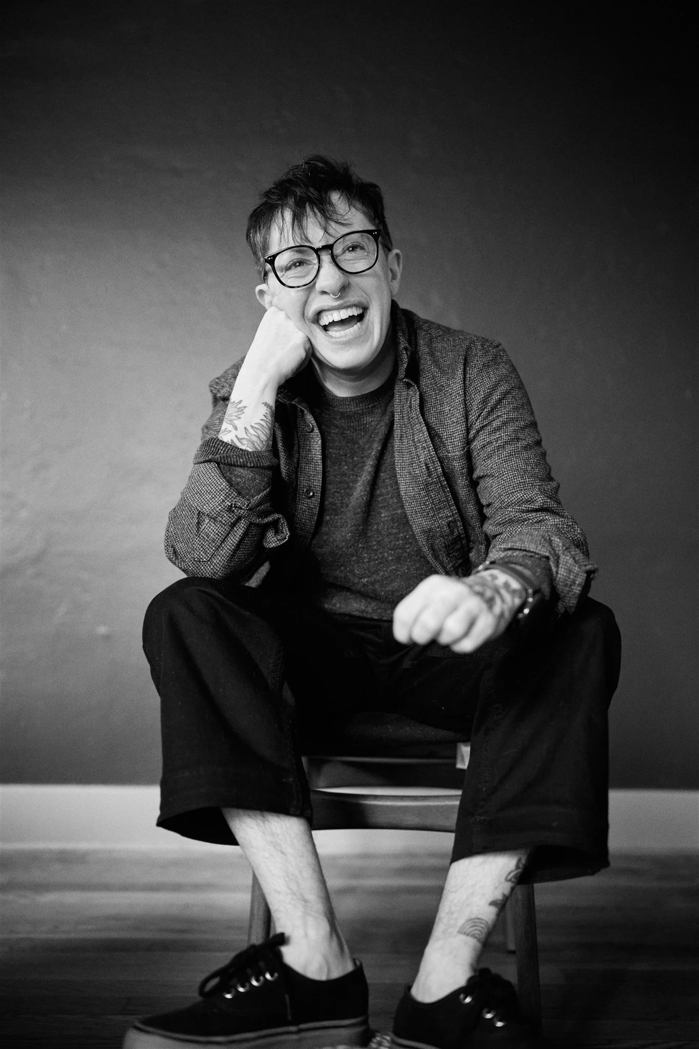 A young man with tattoos on his arms, wearing glasses, a nose ring, a hoodie, and sneakers, sitting on a chair with a dark background, smiling and laughing.