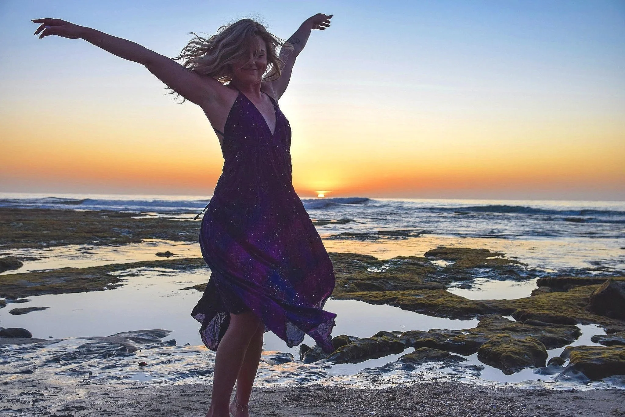 A woman joyfully dancing on the rocks at the beach during sunset.