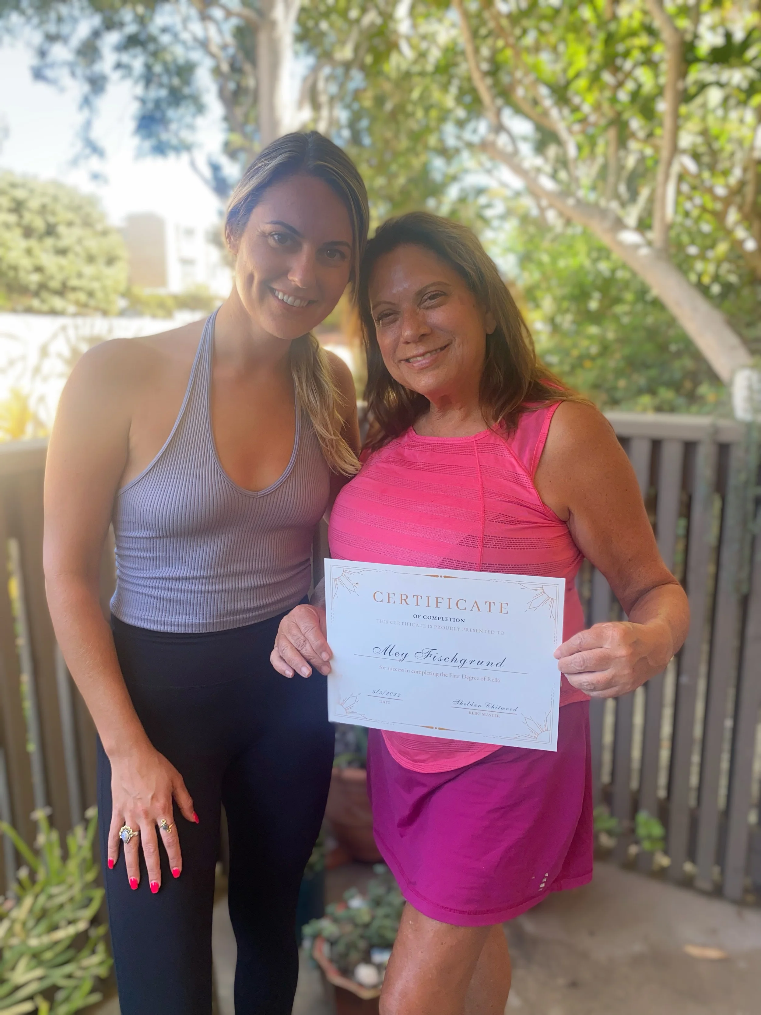 Two women standing outdoors on a wooden deck, one holding a certificate and smiling at the camera. The background is blurred with trees and blue sky.