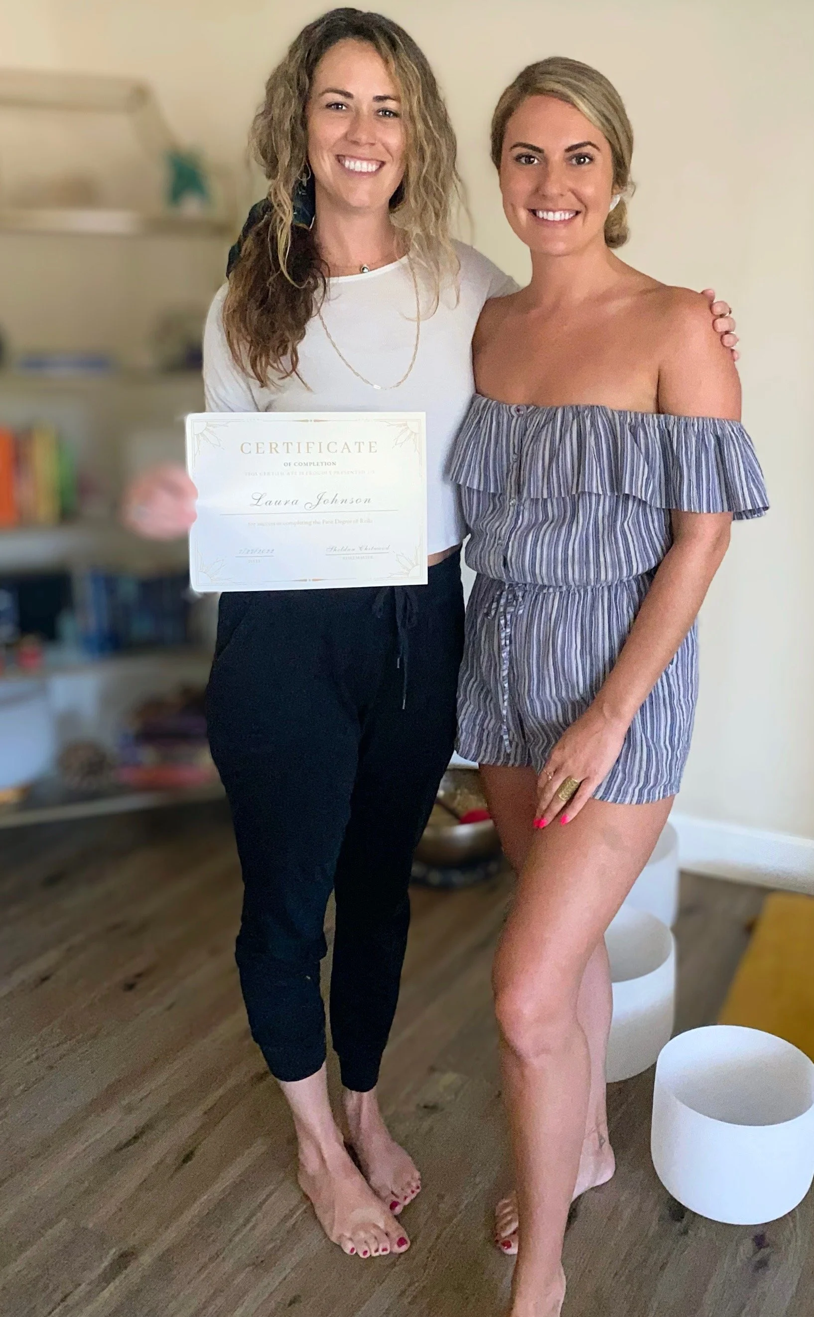 Two women standing close together indoors, smiling at the camera. The woman on the left is holding a certificate, has curly hair, and is wearing a white top and black pants. The woman on the right has blonde hair, is wearing an off-the-shoulder strip