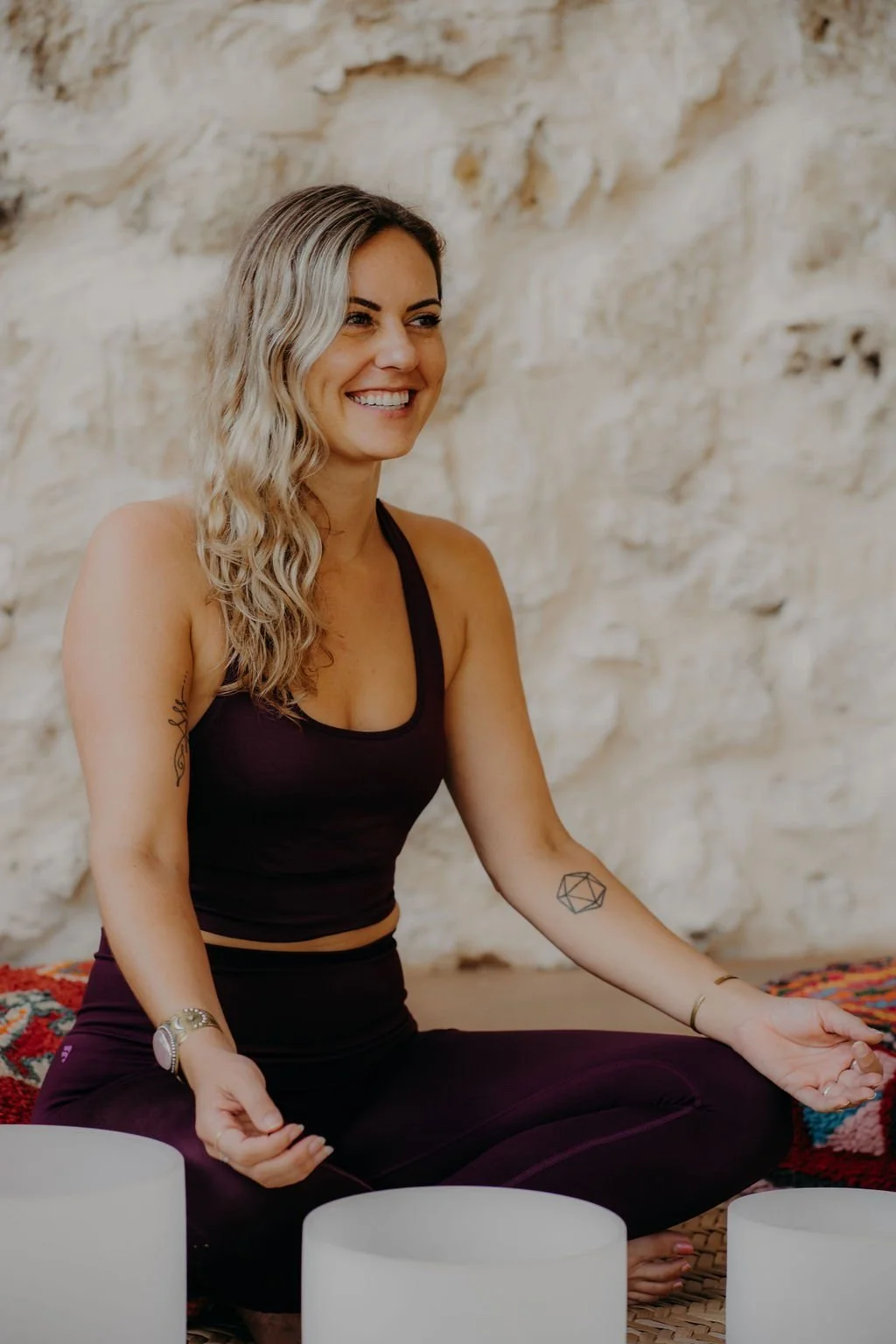 A woman sitting cross-legged on a patterned rug, smiling, engaging in a meditation with white singing bowls in front of her against a textured beige stone wall.