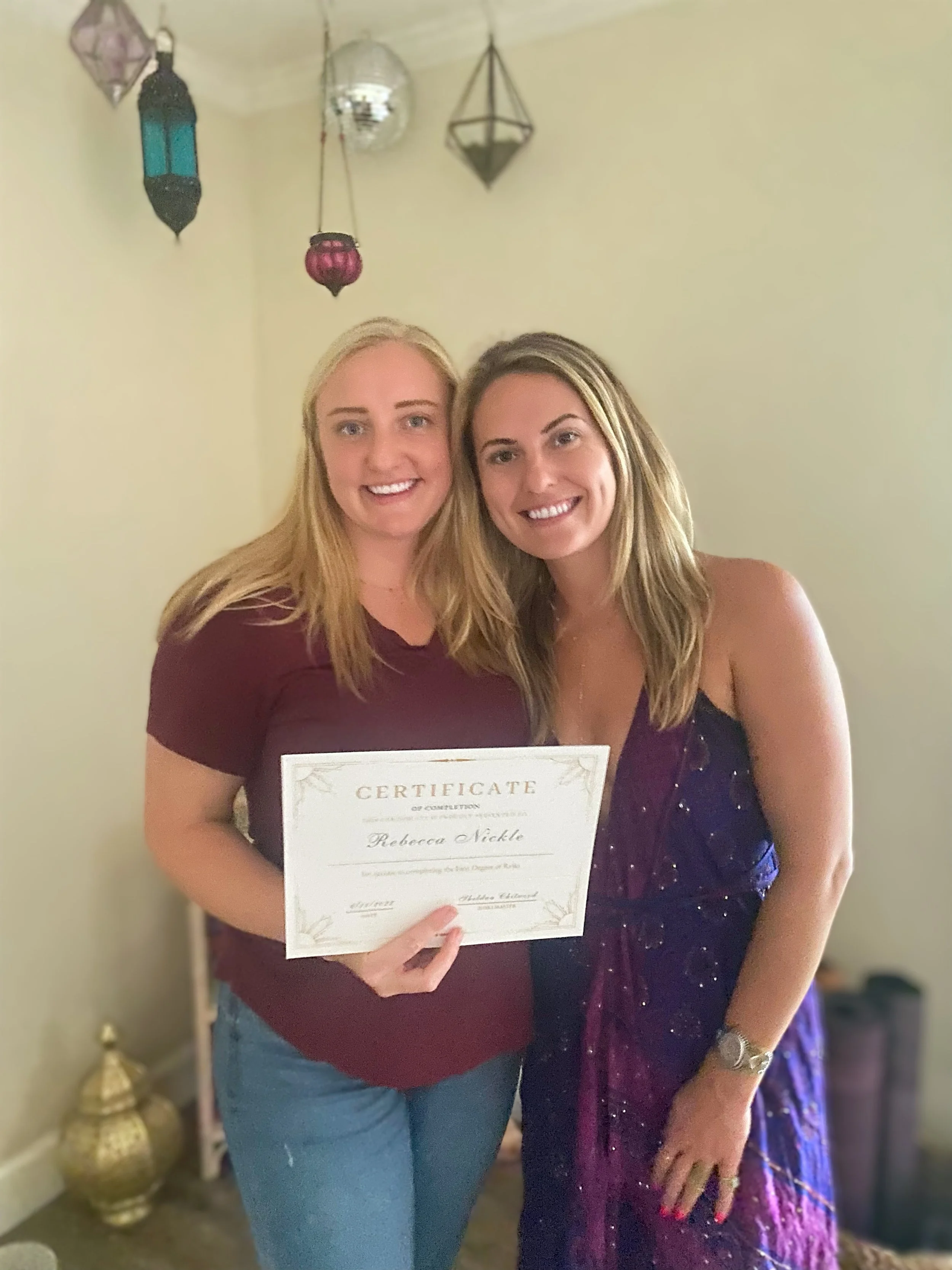 Two women smiling and holding a certificate, standing indoors with decorative hanging lanterns on the wall behind them.