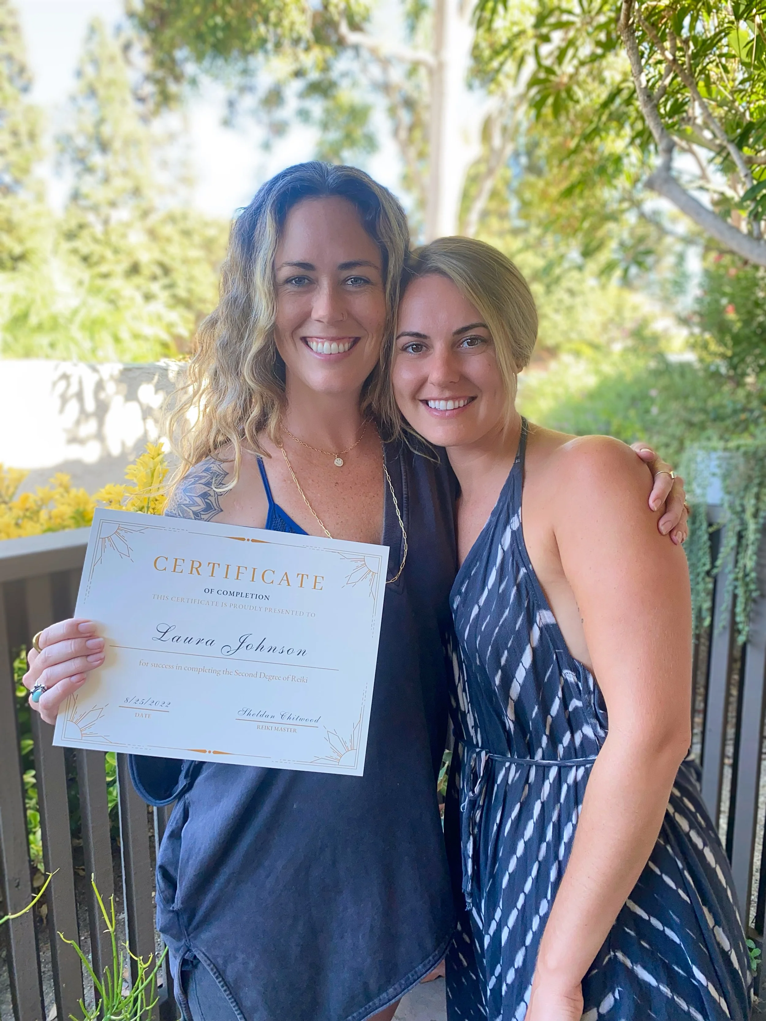 Two women smiling closely together outdoors, one holding a certificate, with greenery and trees in the background.
