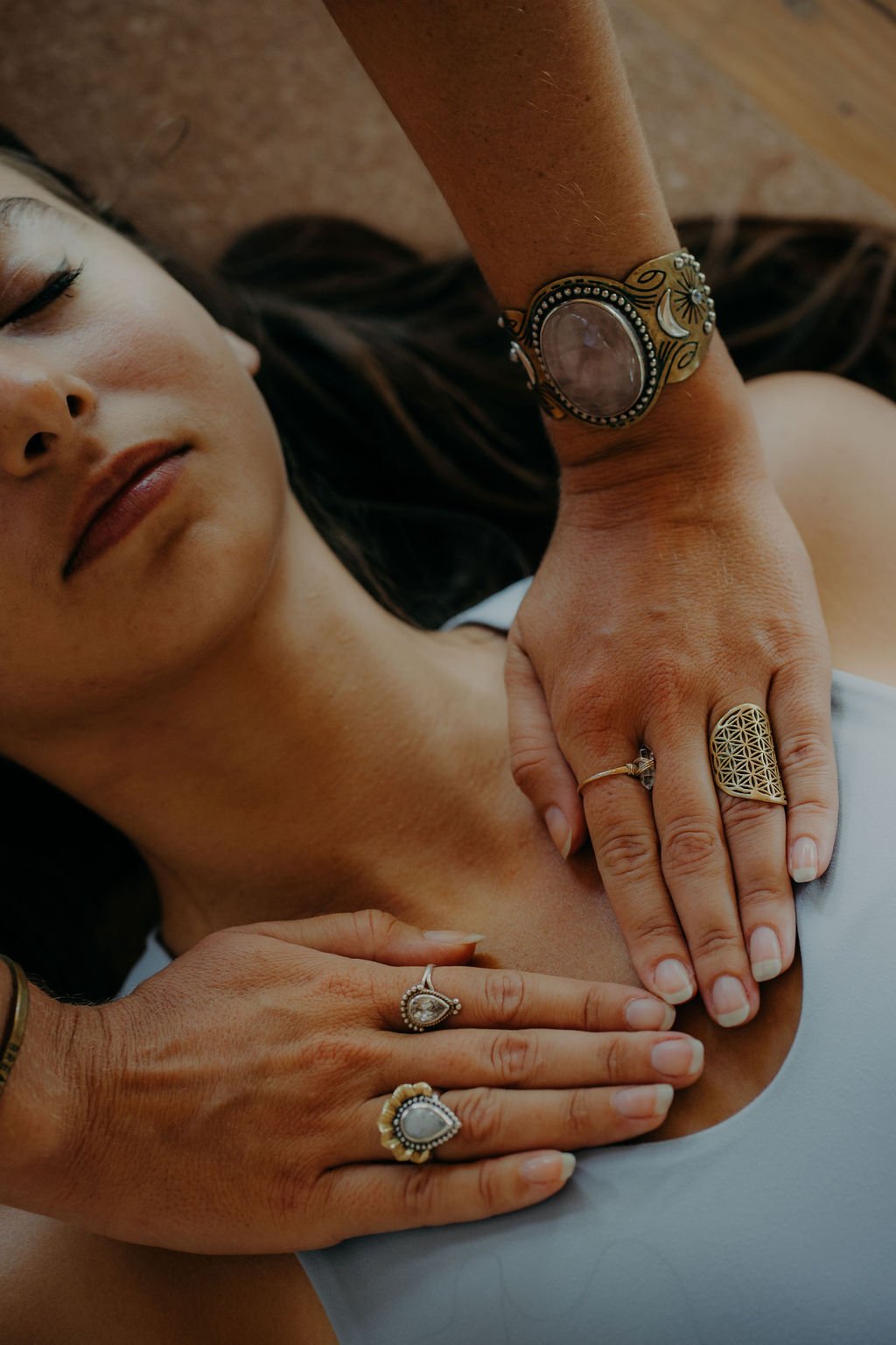 A woman with long dark hair lying down with her eyes closed, wearing rings and a large bracelet, and two hands placed on her chest.