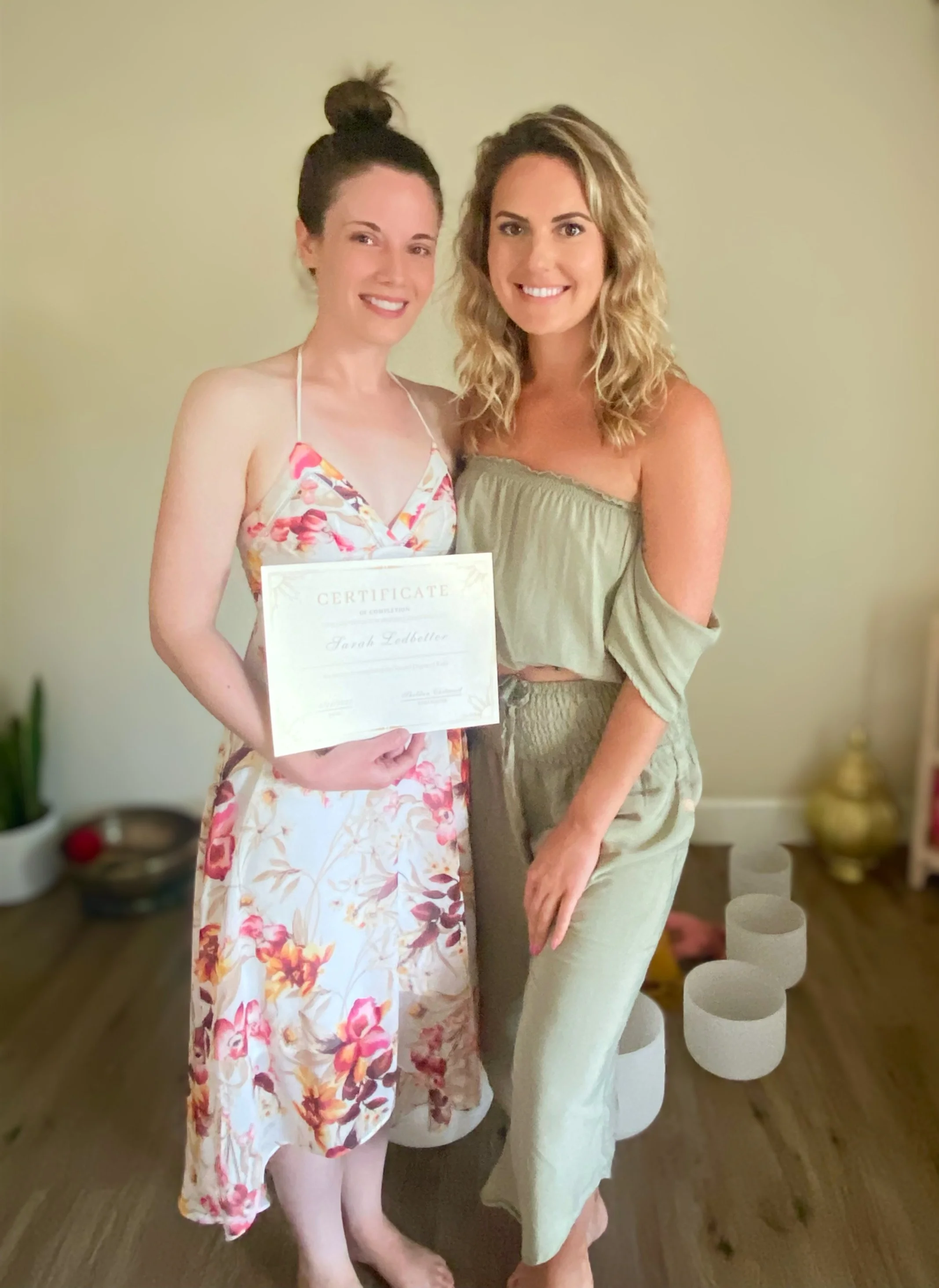 Two smiling women standing together indoors, one holding a certificate, both dressed in casual summer clothing, with a neutral-colored wall and home decor in the background.