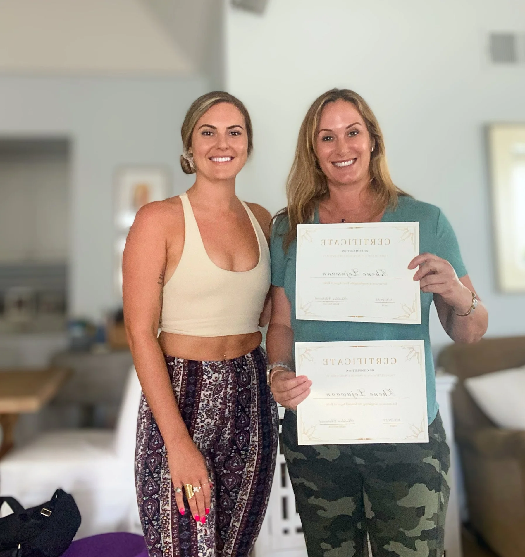 Two women standing together, smiling, holding certificates, one woman wearing a cream-colored top and patterned pants, the other wearing a teal top and camouflage pants.