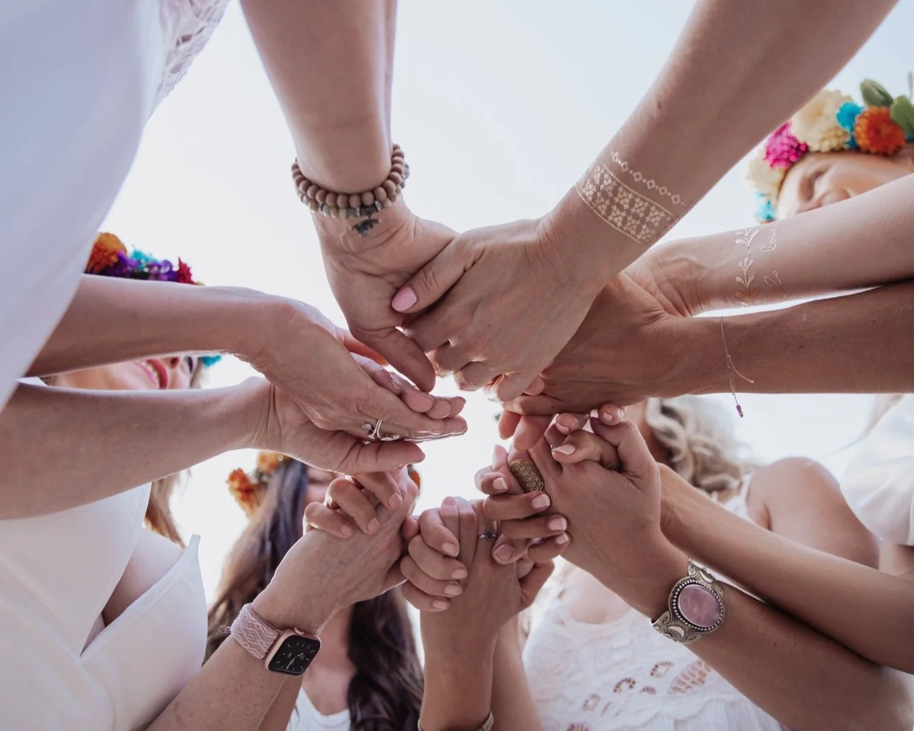 Group of women with hands intertwined, participating in a unity or friendship gesture, with some wearing bracelets, rings, and tattoos.