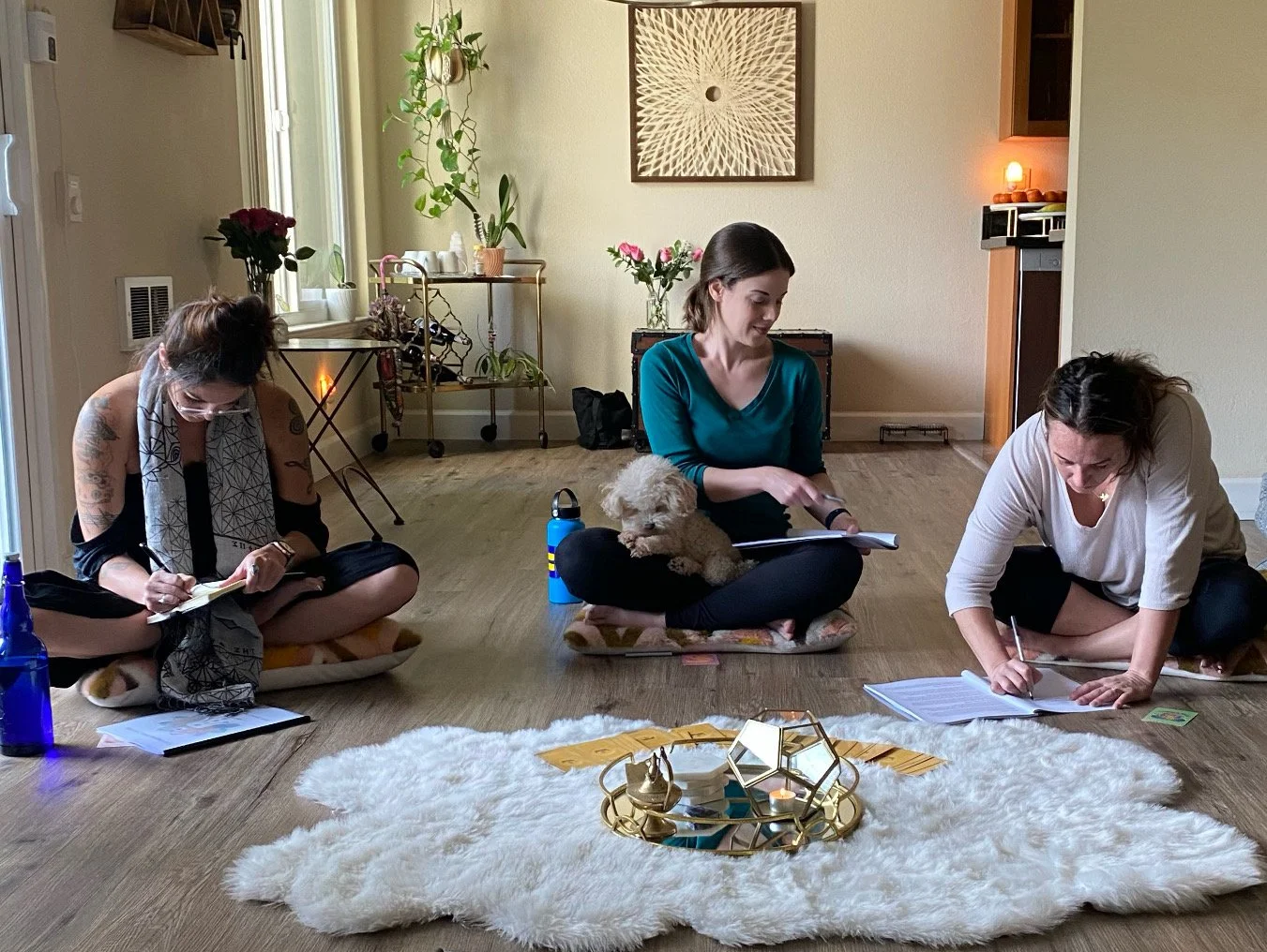 Three women sitting on the floor in a casual home setting, writing or reading, with a small white dog on one woman's lap, surrounded by notebooks, papers, a blue water bottle, and a decorative candle holder on a white fluffy rug, with background plan