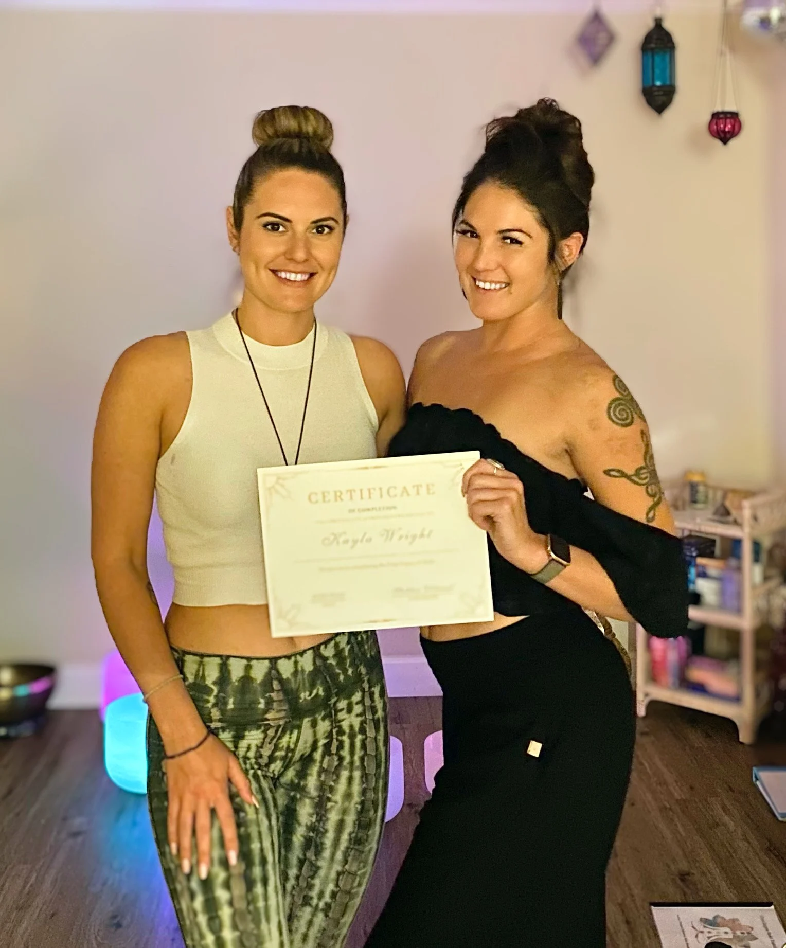 Two women standing side by side, smiling, with one holding a certificate. The woman on the left has her hair in a bun, wearing a sleeveless crop top and patterned pants. The woman on the right has dark hair in a bun, wearing an off-shoulder black dre