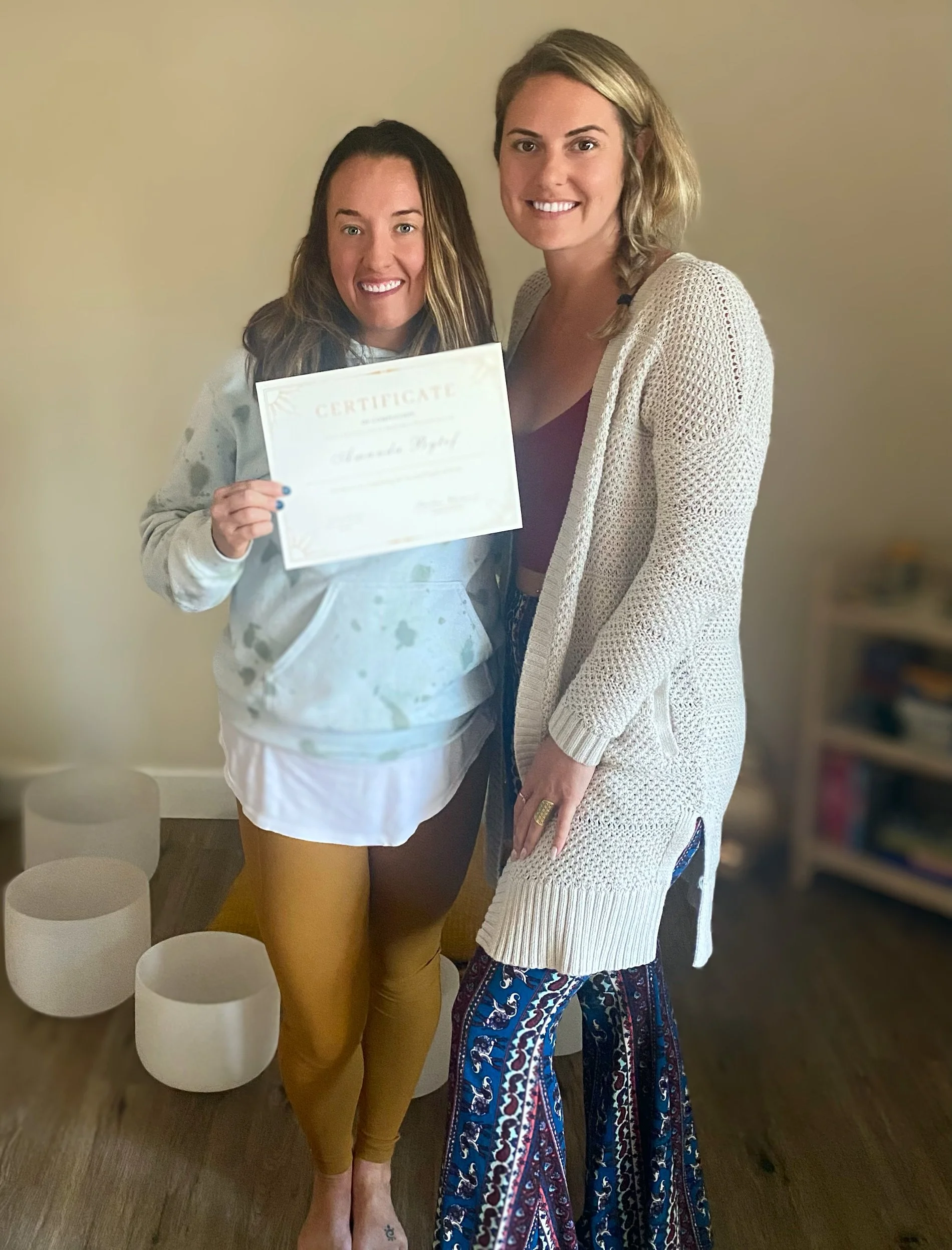 Two women standing together, one holding a certificate, smiling for a photo indoors. The woman on the left has dark hair and light skin, wearing a white hoodie and mustard pants. The woman on the right has blonde hair, light skin, wearing a cream car
