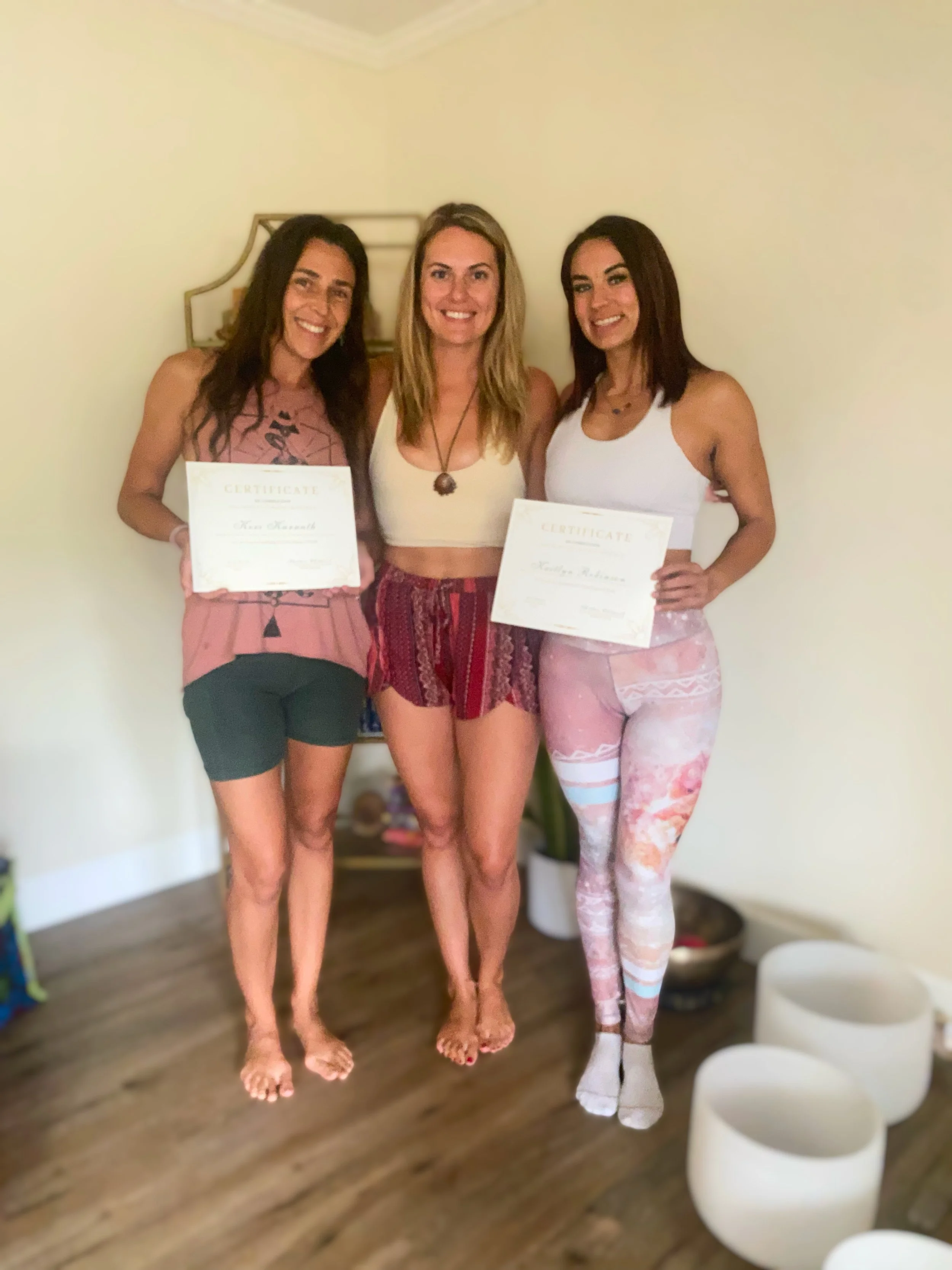 Three women standing indoors, smiling, holding certificates, with a beige wall and a small mirror behind them. They are dressed in casual summer clothing.