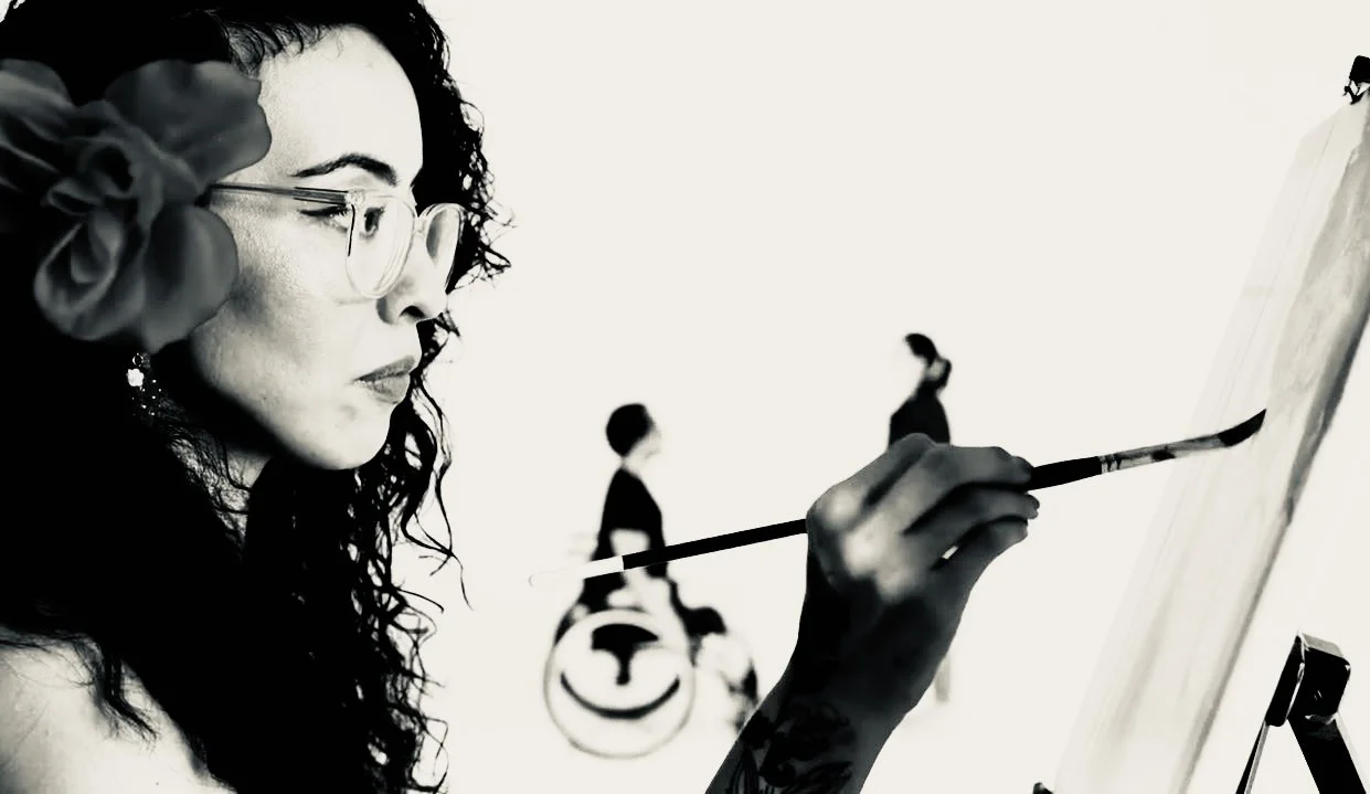 Artist Genevieve Ramos with curly hair and glasses painting a mural featuring an interable couple in the distance.