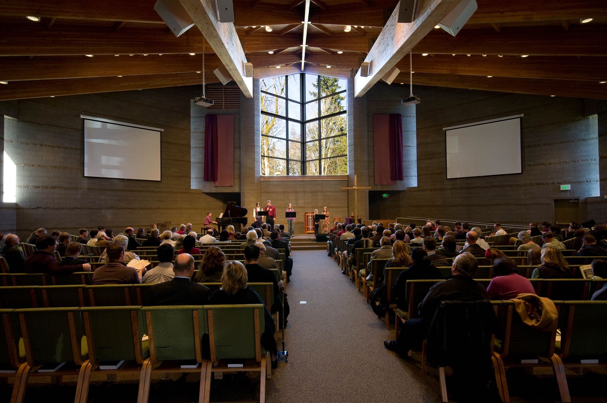 Congregation attending a church service or concert inside a modern church building with large stained glass window, wooden ceiling, and two large projection screens.