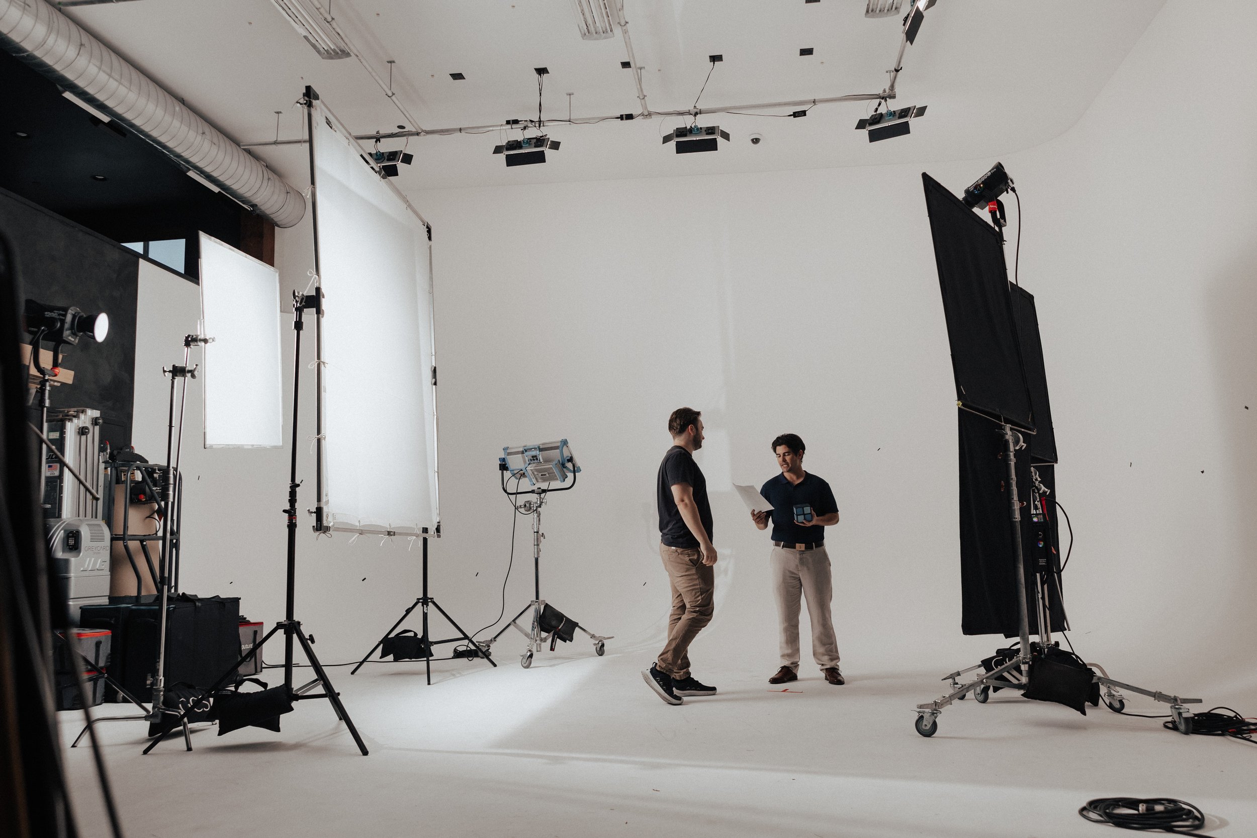 Two men standing in a photography or video studio surrounded by lighting and camera equipment, with a white backdrop and a ceiling with hanging lights and pipes.