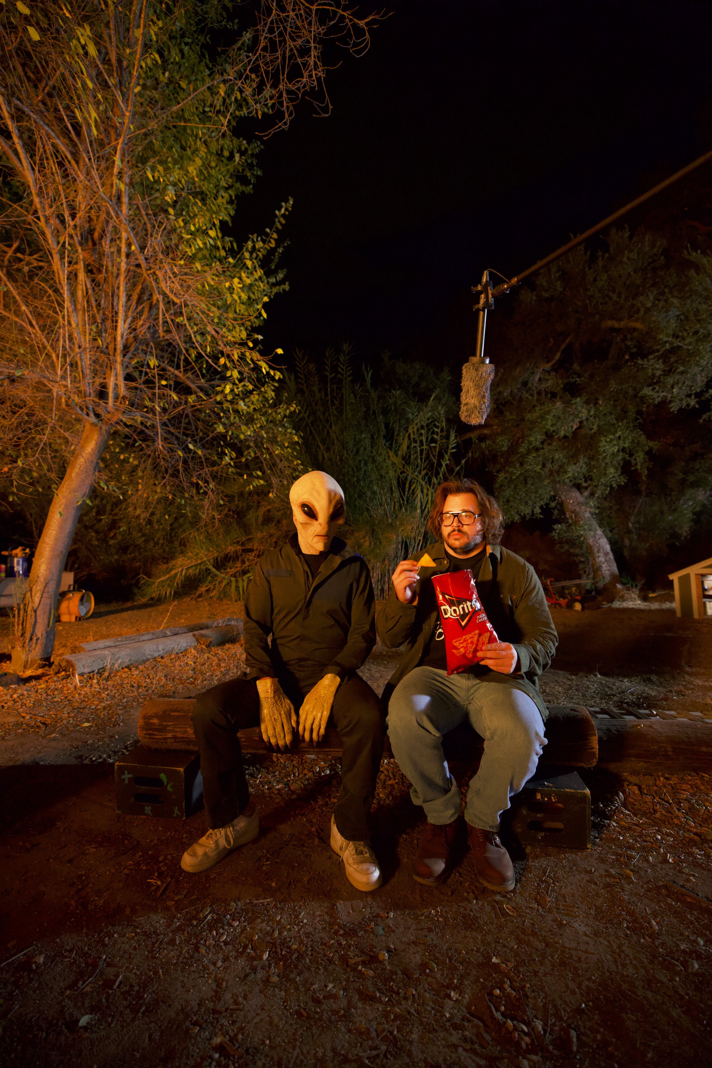 A person in a creature costume sitting on a bench next to a man eating chips, outdoors at night, with trees and filming equipment overhead.