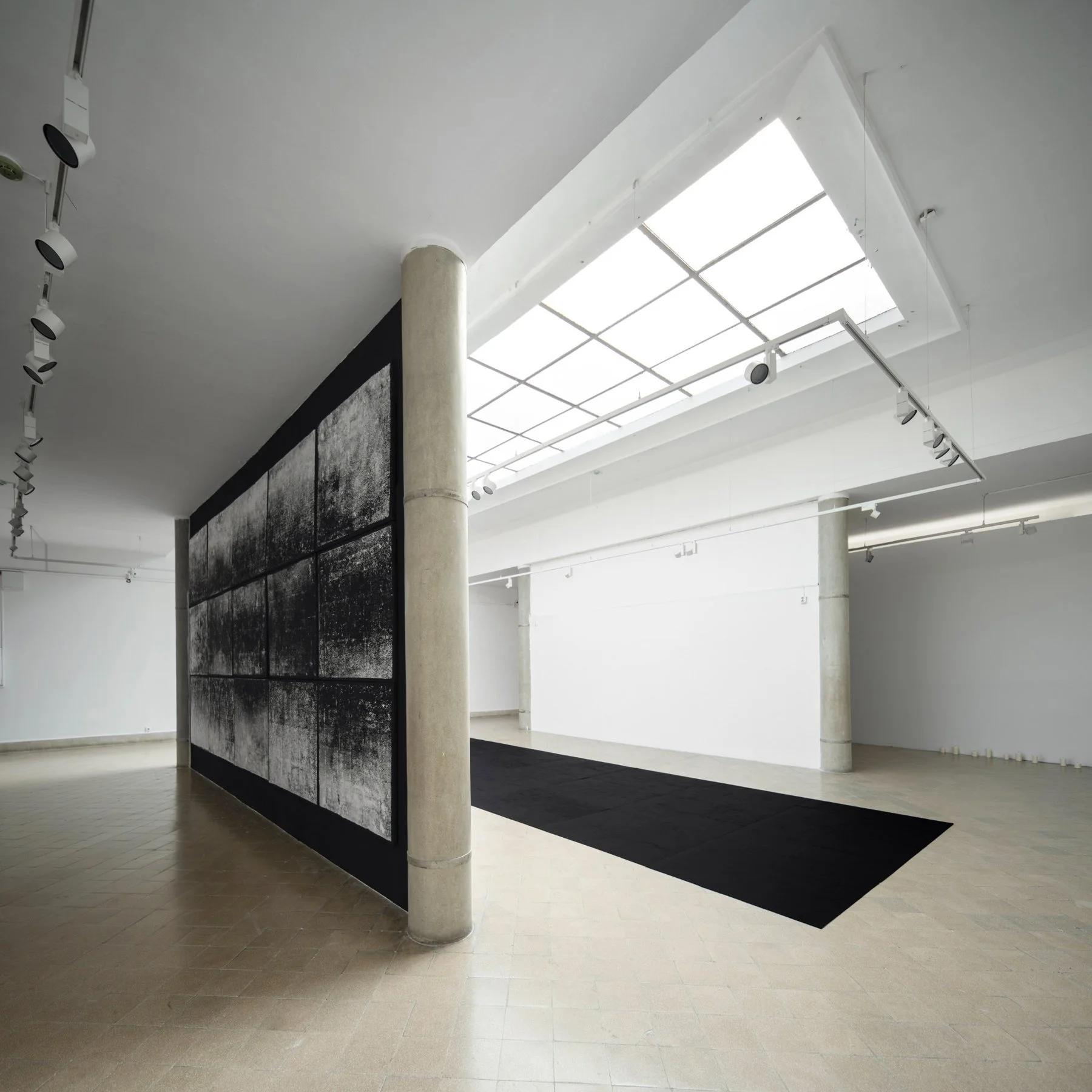 Oblique angle of Koós’s light-absorbing installation, with wall-mounted prints and black floor alignment under the skylight.