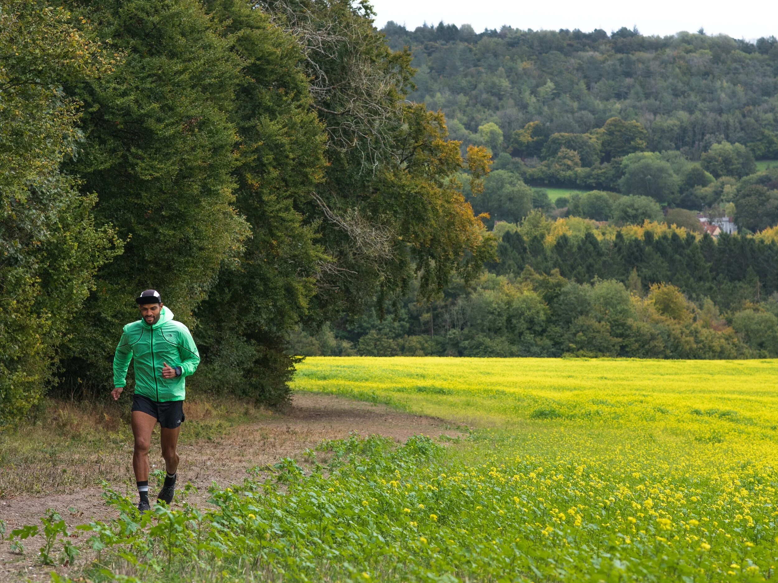 A man running along a dirt trail through a lush green field with yellow wildflowers, surrounded by dense trees and rolling hills in the background.