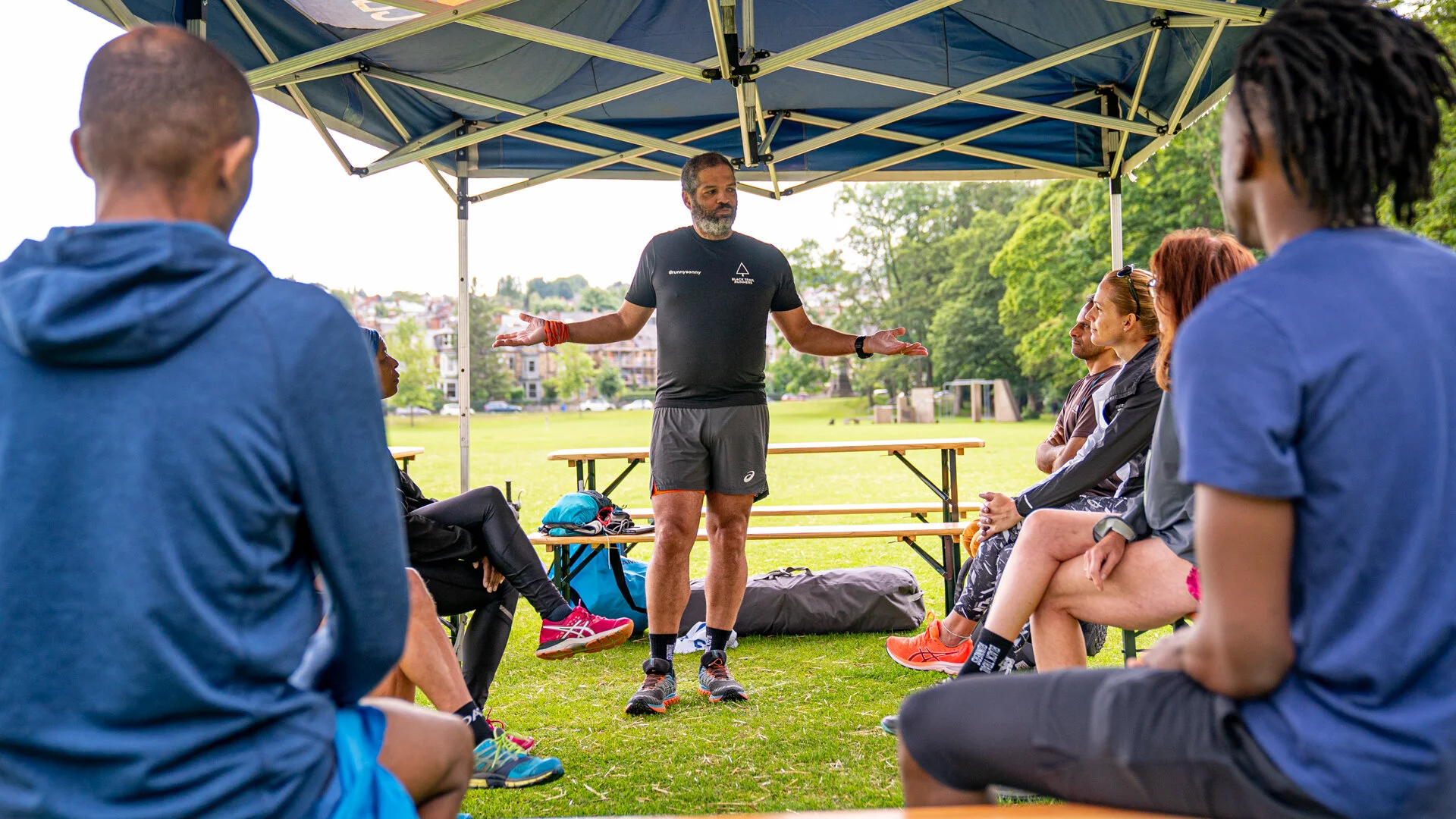 A group of people sitting outdoors under a canopy, listening to a man in athletic attire who appears to be leading a discussion or giving instructions.