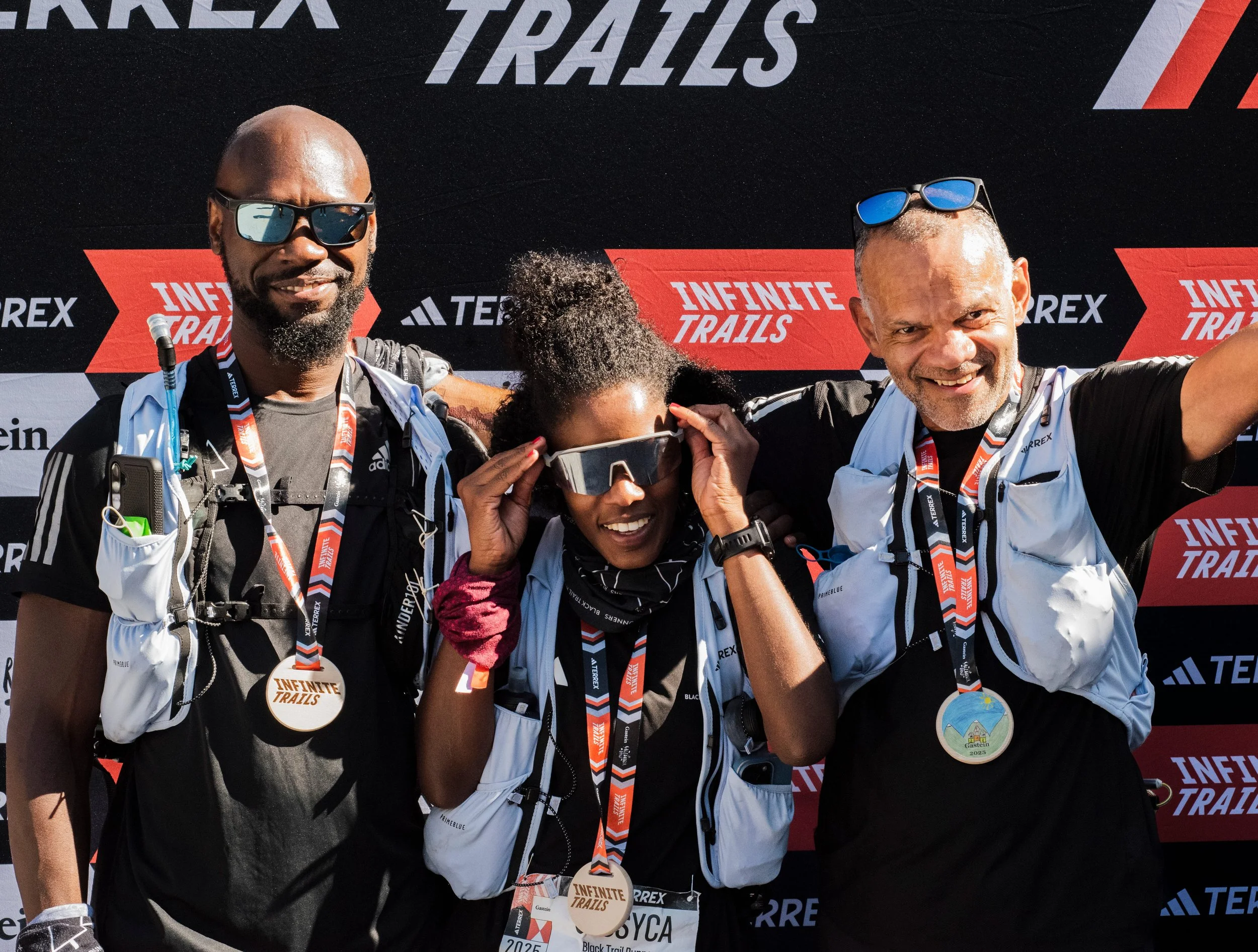 Group of people celebrating and smiling at an outdoor trail running race event with a large black and red banner that has the words 'Infinite Trails' on it, wearing medals and athletic gear.