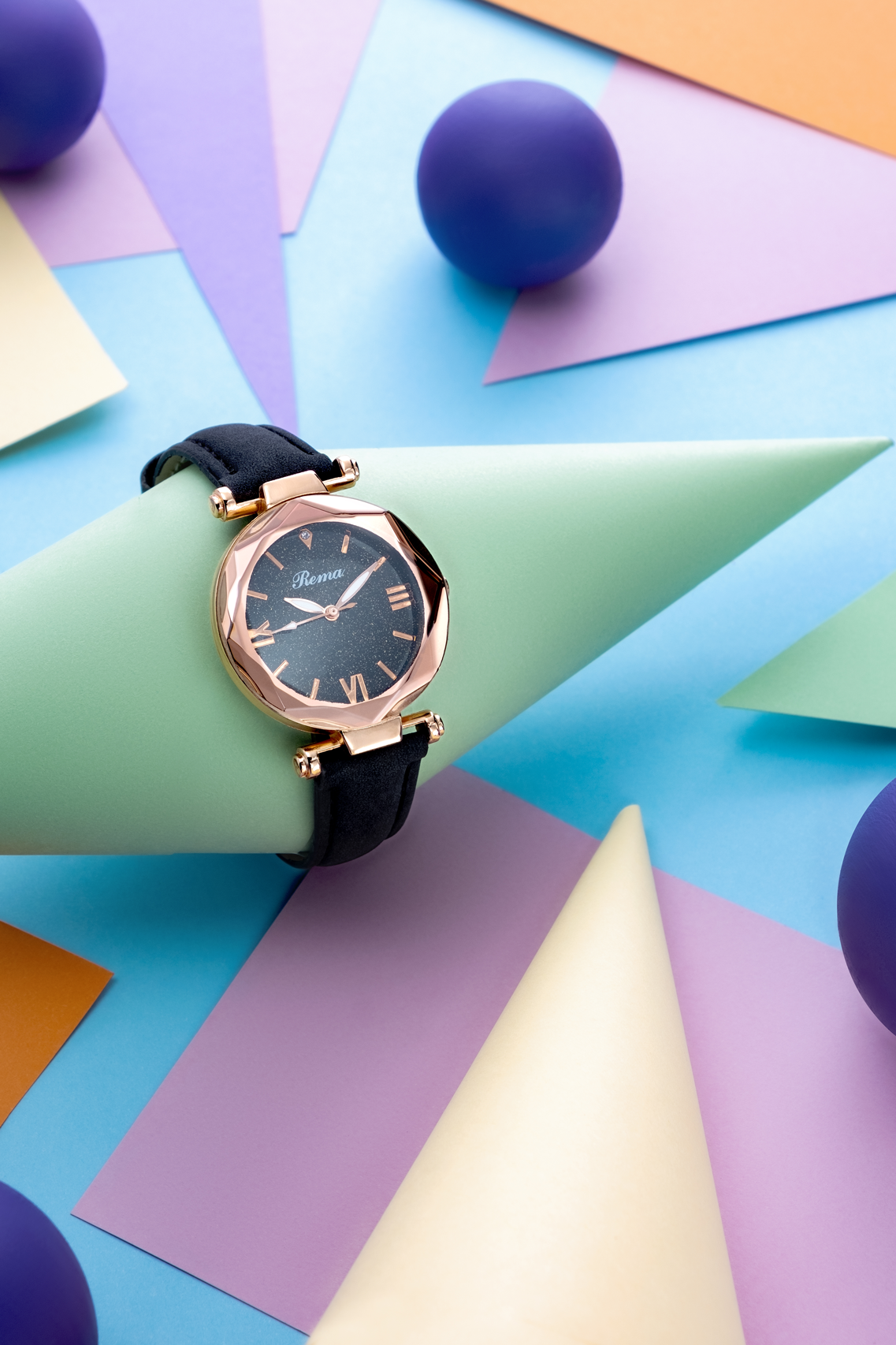 A black and gold wristwatch resting on a green paper cone, surrounded by colorful paper sheets and purple paper balls on a pastel background.