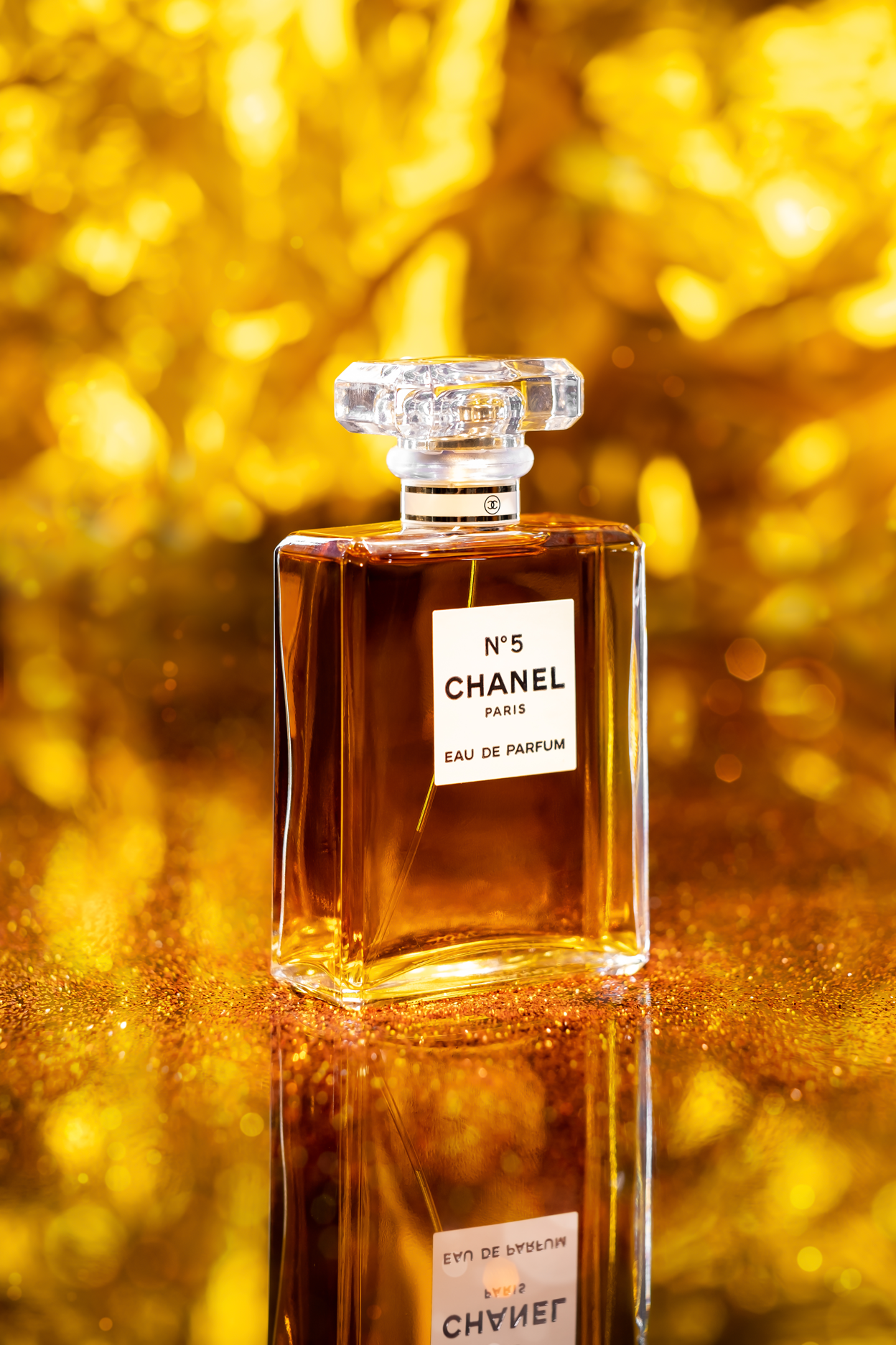 A bottle of Chanel No. 5 perfume against a golden, glittery background with a reflection on a shiny surface.