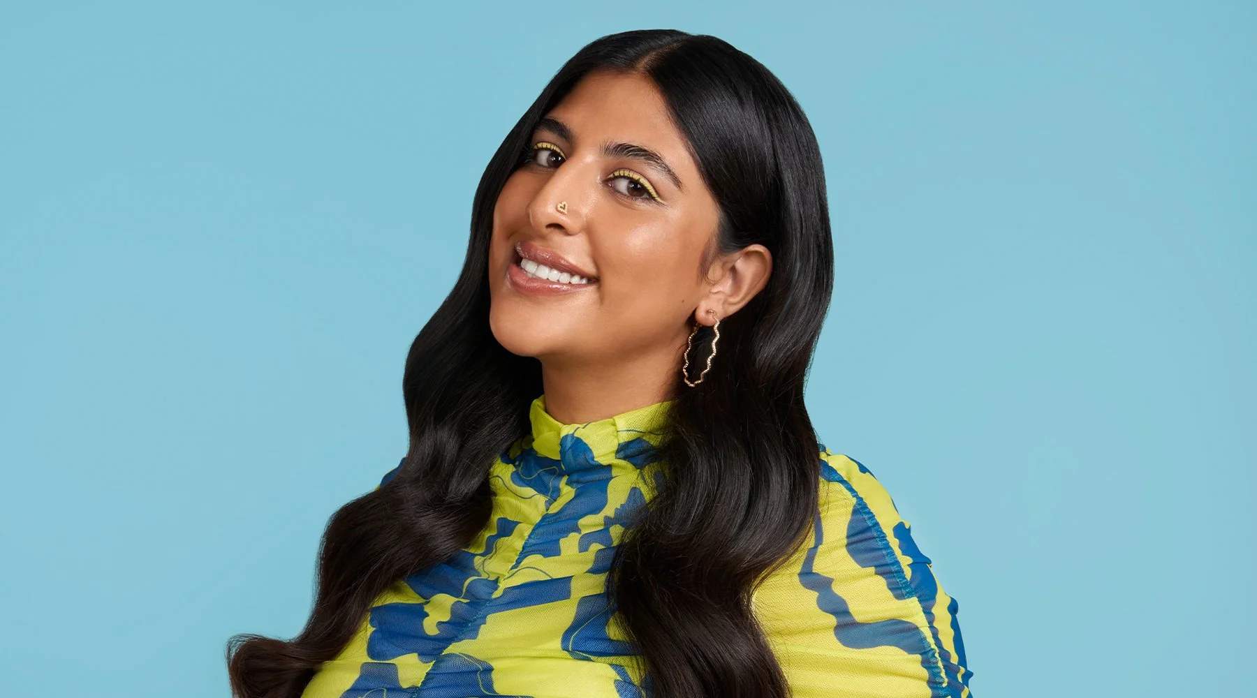 Smiling South Asian heritage model demonstrating Noughty Haircare flyaway wand on colourful blue background