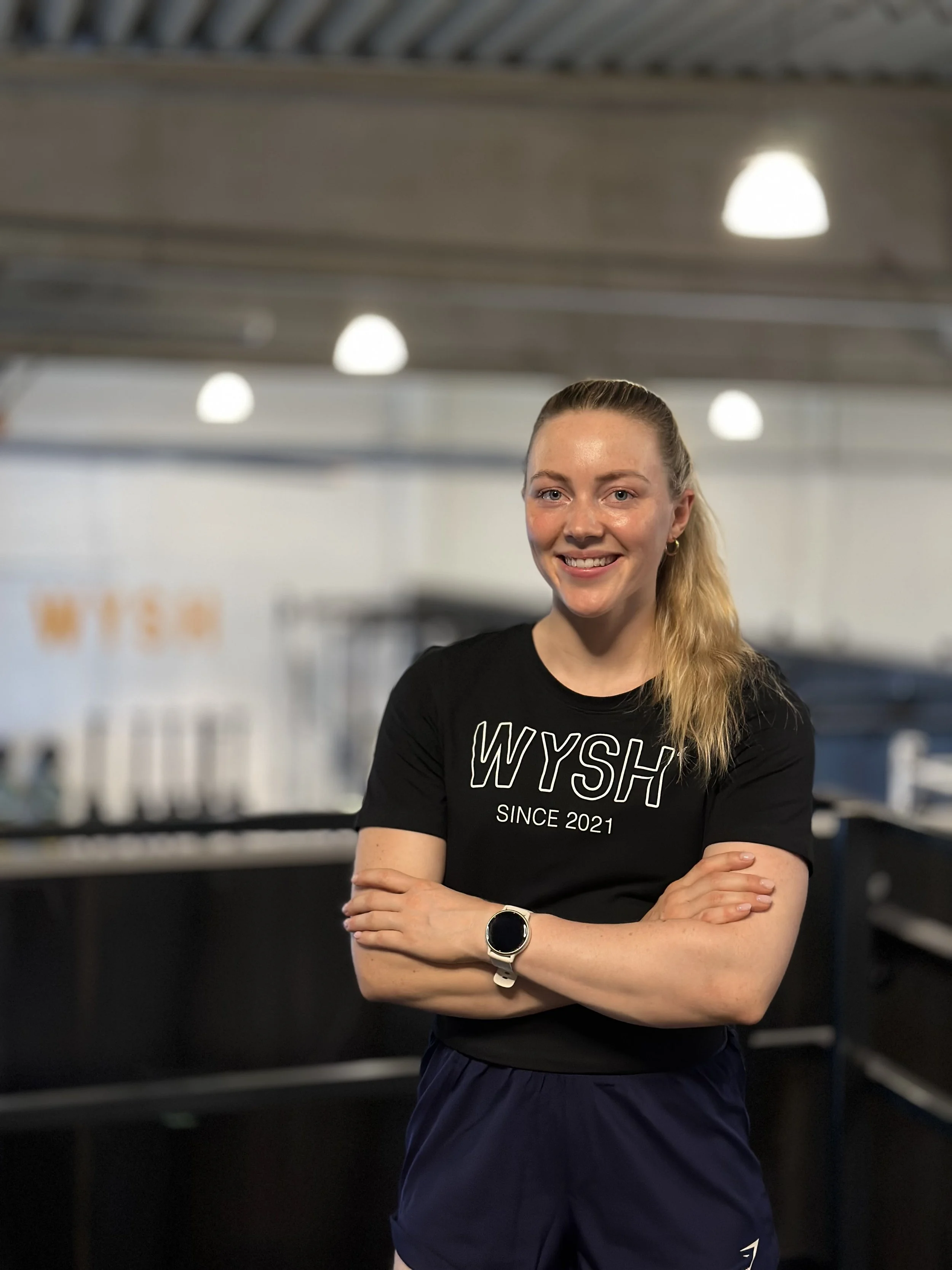 Trainerin Elena Smeenk, eine junge, lächelnde Frau mit blondem Haar trägt ein schwarzes T-Shirt mit dem Schriftzug 'WYSH SINCE 2021' und steht in dem Fitnessstudio WYSH.Fitness, mit verschränkten Armen.