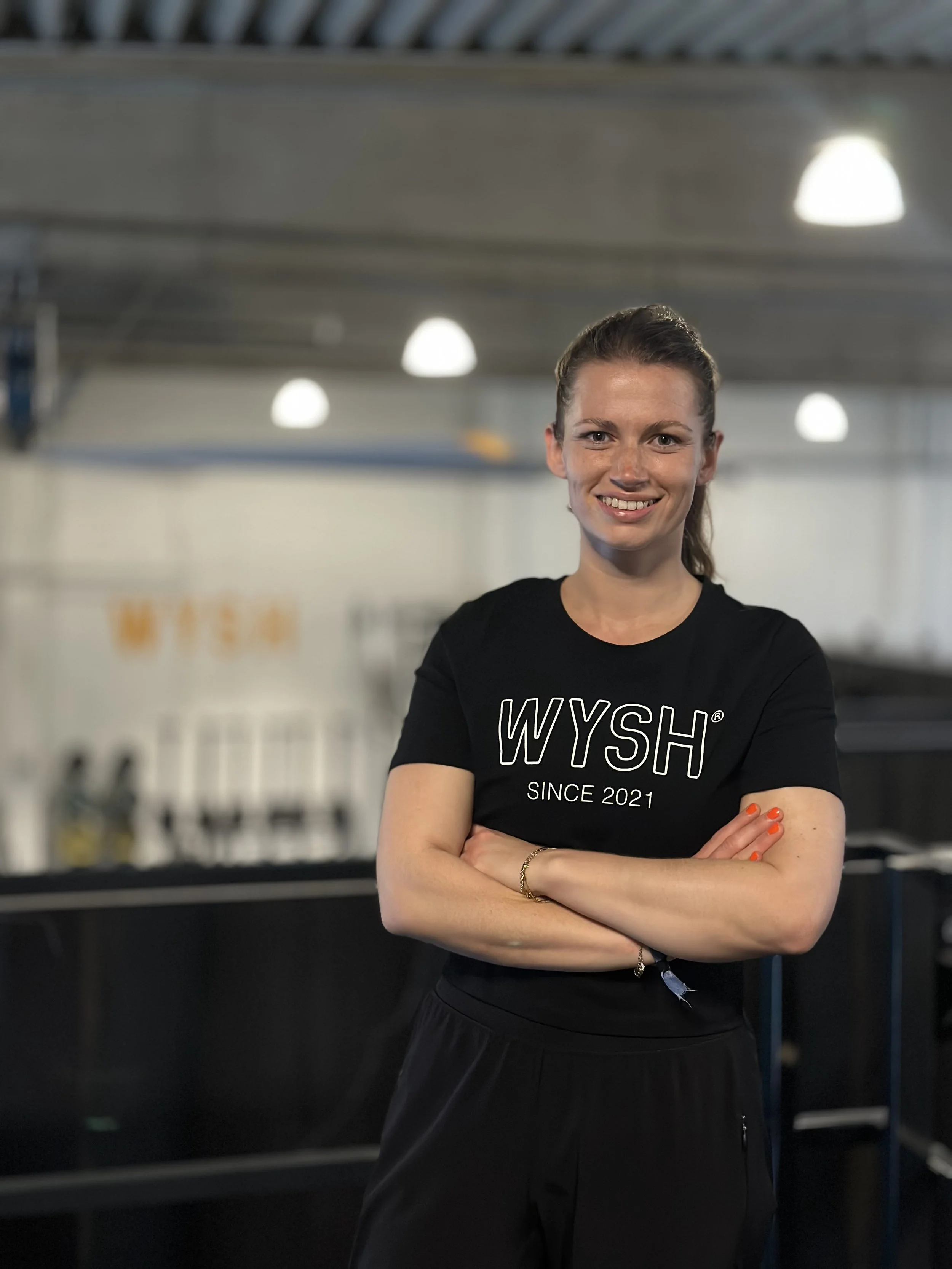 Trainerin Lea Roosen, eine lächelnde, junge Frau trägt ein schwarzes T-Shirt mit der Aufschrift "WYSH SINCE 2021" und steht in dem Fitnessstudio WYSH.Fitness.