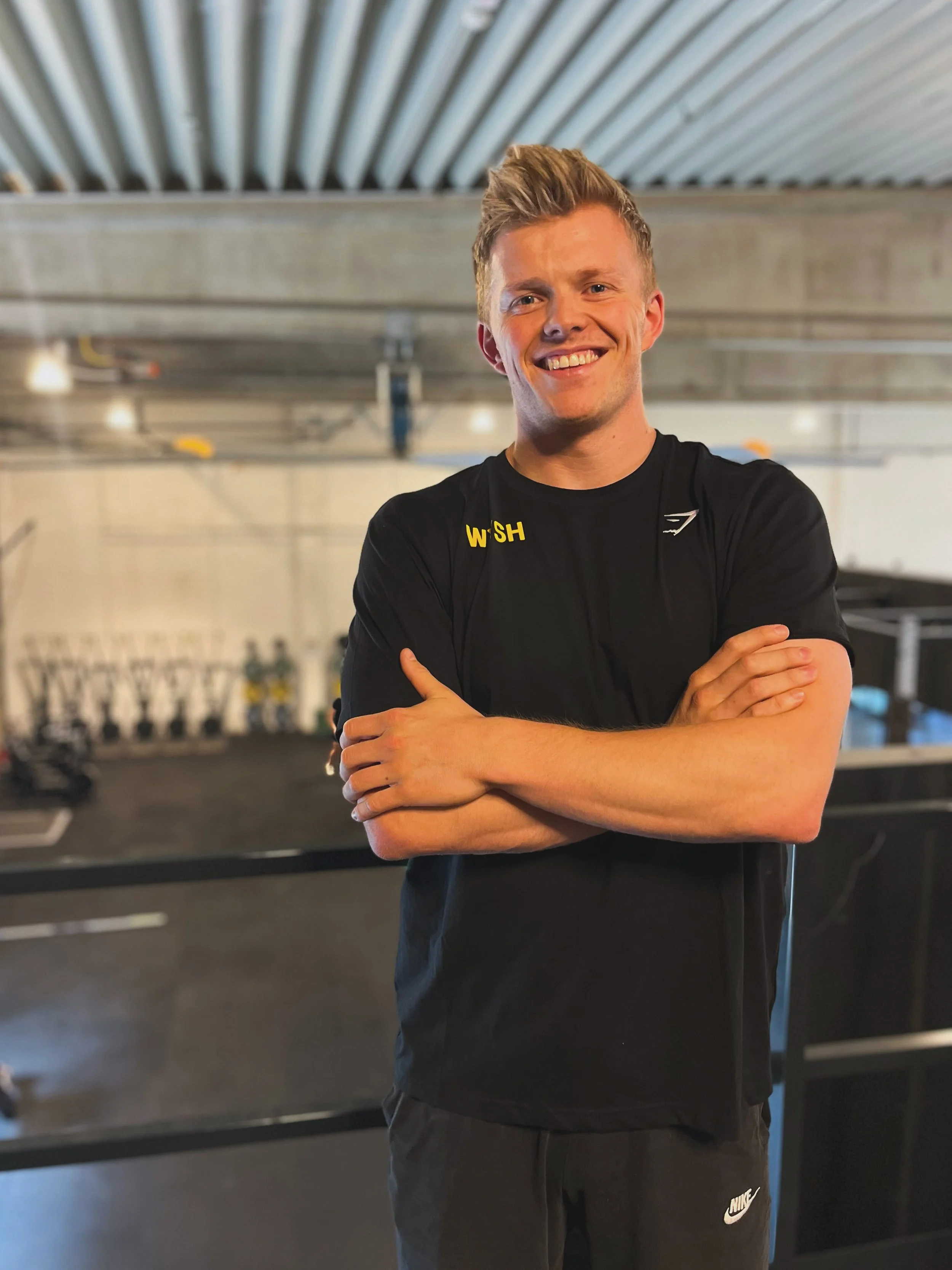 Trainer Moritz Runge, ein lächelnder, junger Mann in schwarzem Sportshirt, steht mit verschränkten Armen in dem Fitnessstudio WYSH.Fitness.