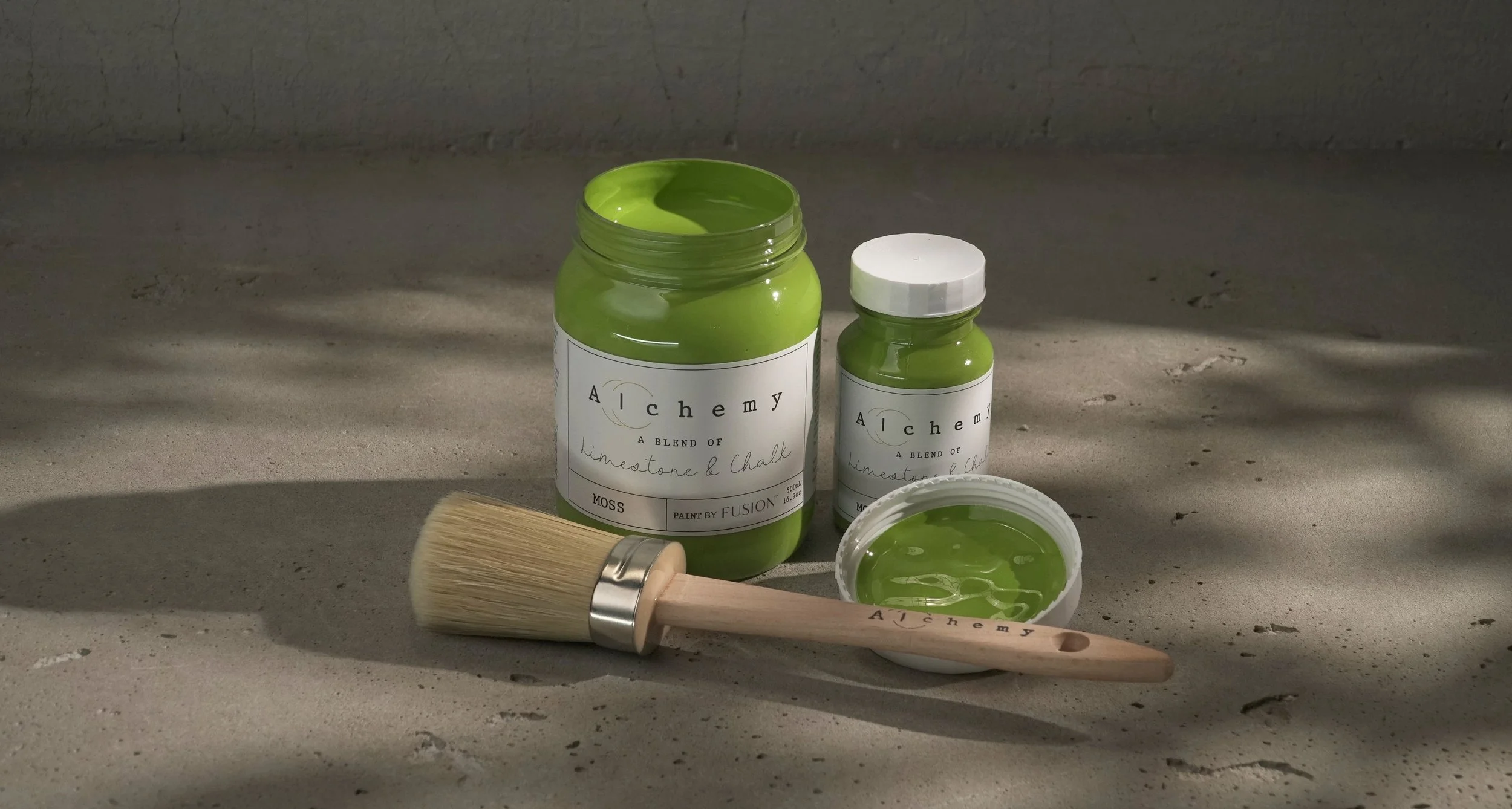 Open jar of green paint labeled 'Alchemy', smaller jar of matching paint, paintbrush with green paint, and a small container of green paint on gray surface.
