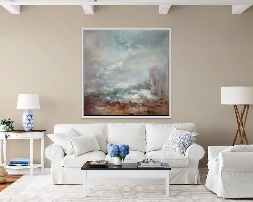 Living room with beige walls, white sofa, abstract landscape painting, white side table with lamp, and blue flowers on coffee table.