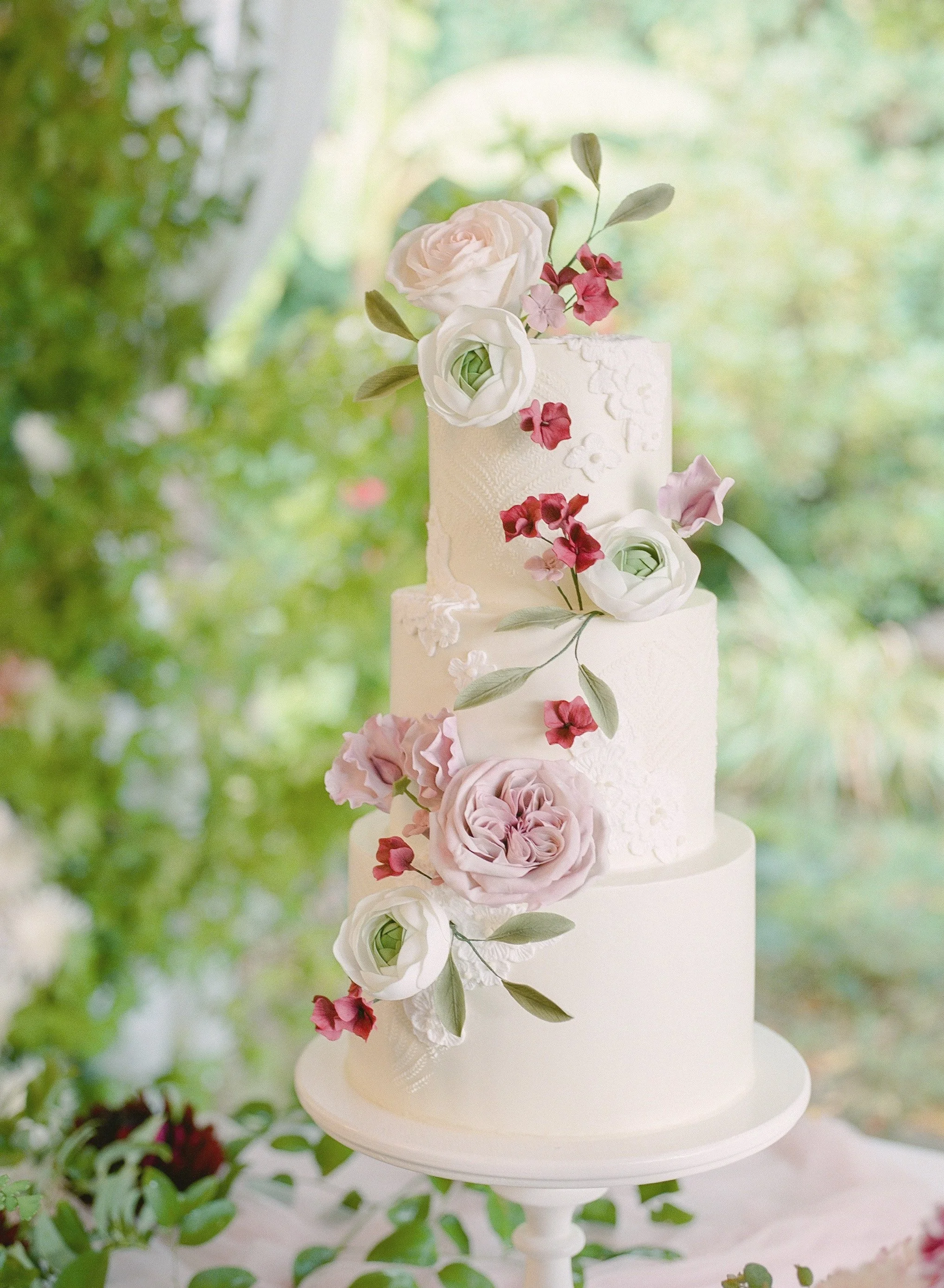 Custom three-tiered wedding cake with Art Deco stencil design and handmade lace appliqués, adorned with handmade sugar flowers in a color palette of plum, pale lavender, and blush pink, with delicate green accents. In a green outdoor setting.