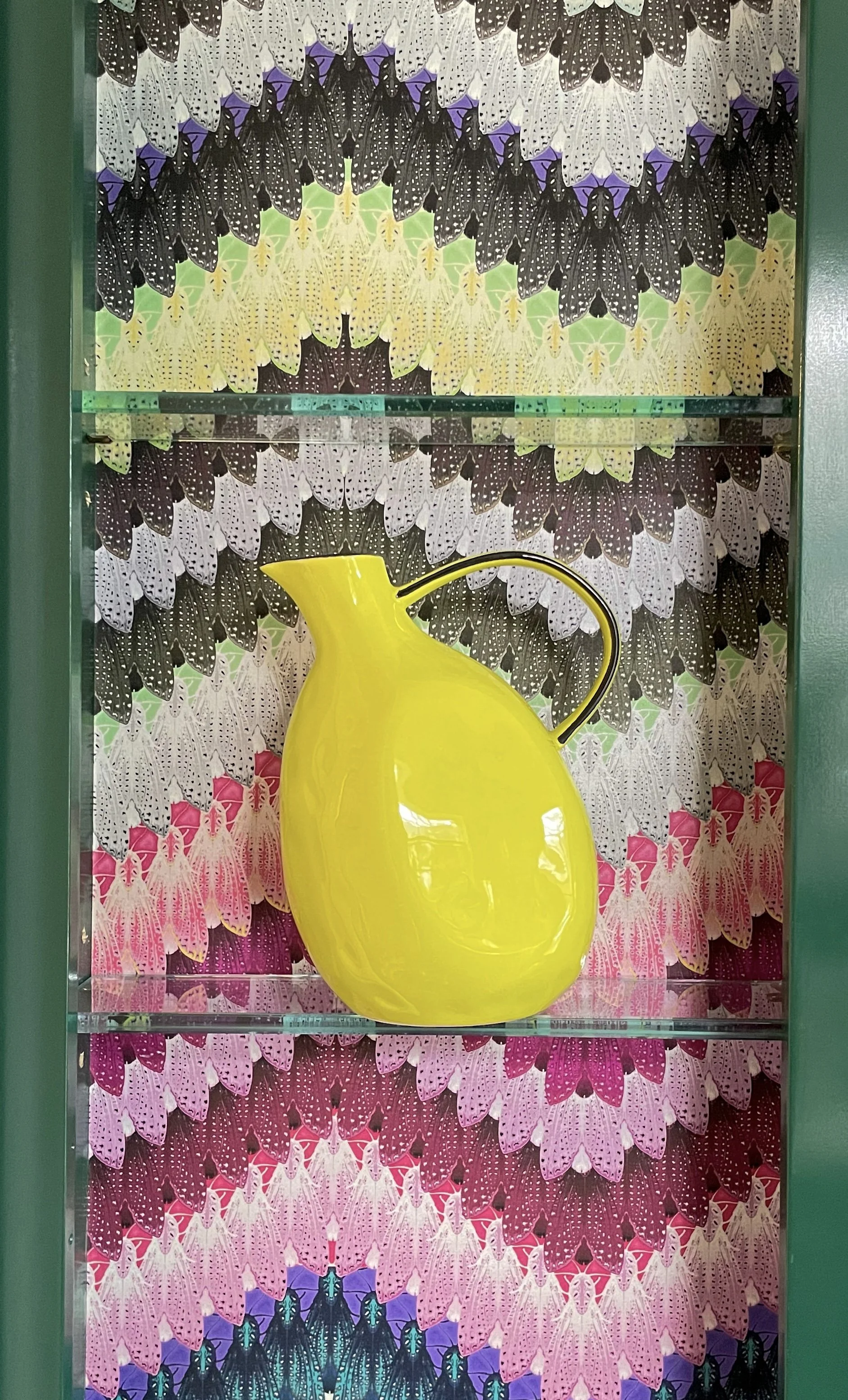A bright yellow ceramic pitcher on a glass shelf, with a butterfly-patterned wallpaper background featuring black, purple, yellow, green, pink, and white butterflies.