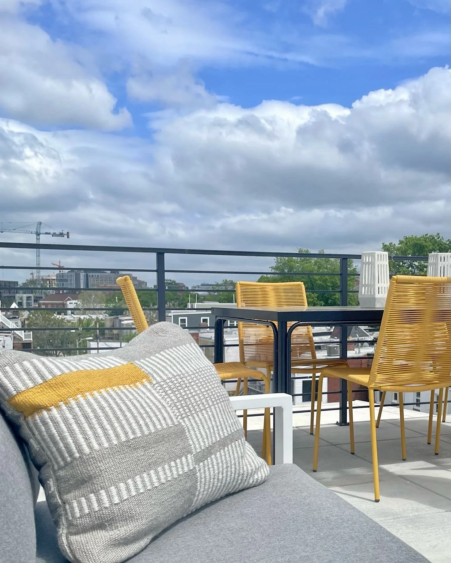 Rooftop patio with a gray couch and colorful cushions, yellow rattan chairs, black metal table, white lanterns, and city view with blue sky and clouds.