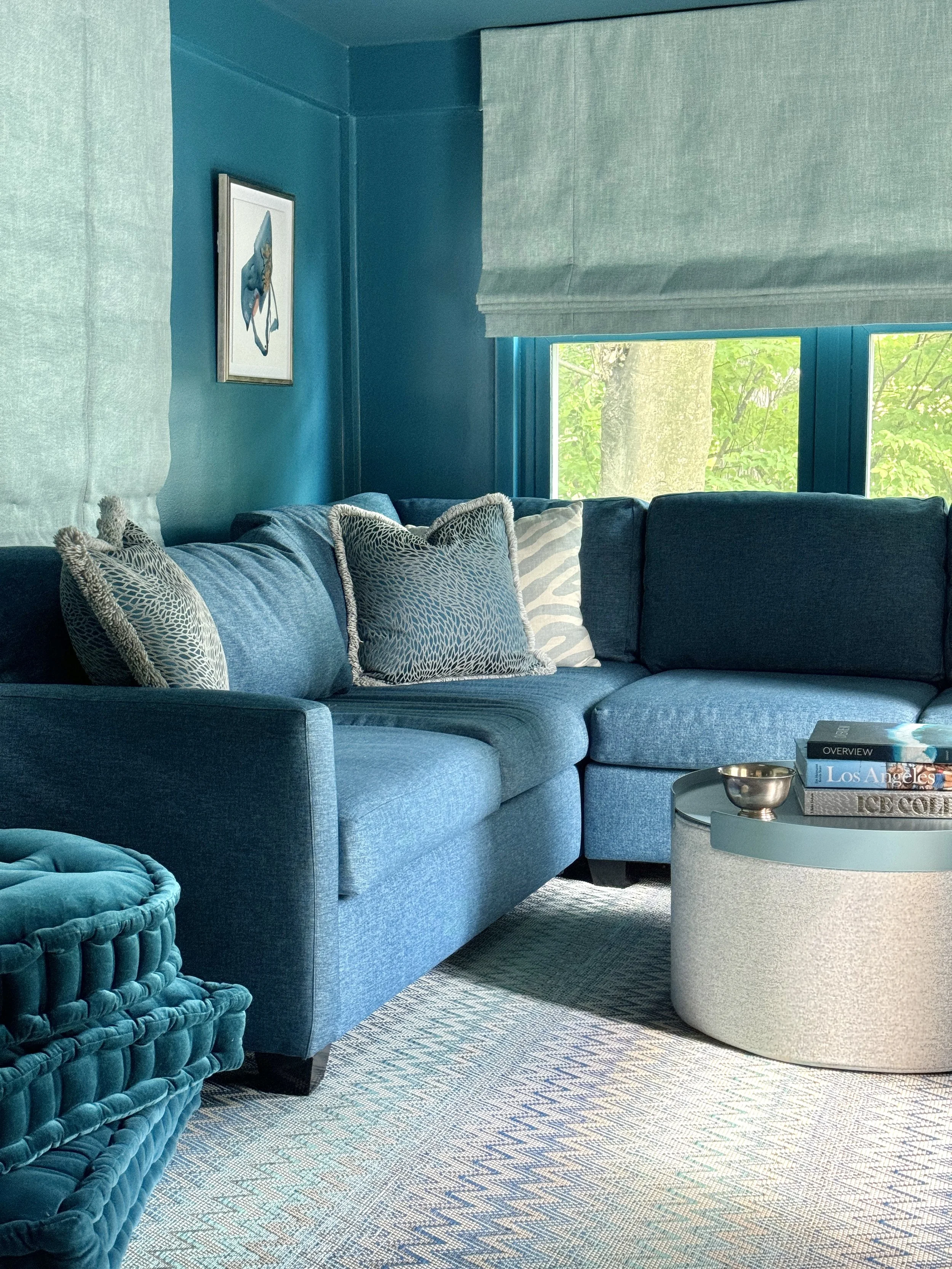 A teal-colored living room with a sectional sofa, patterned pillows, a round coffee table with books, a small metallic bowl, and a window showing green trees outside.
