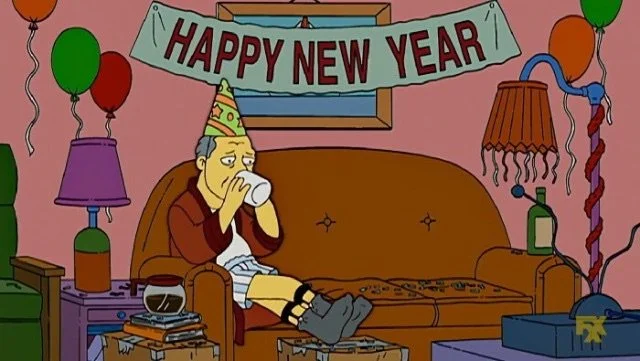screenshot from the cartoon series "The Simpsons" were a man is in a robe and socks on the couch sadly drinking coffee in a dirty post-party home