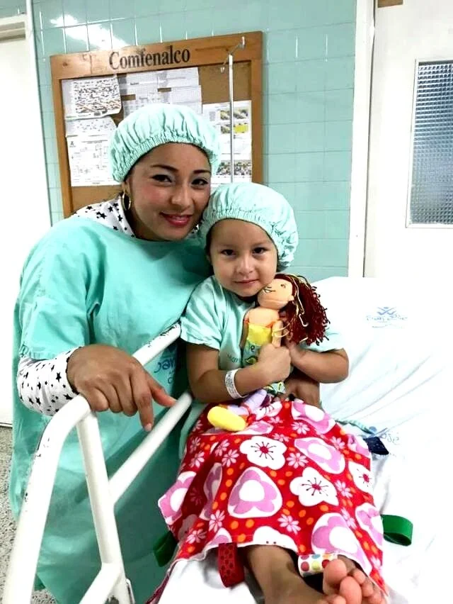 A woman and a young girl sitting on a hospital bed, both wearing green hospital gowns and caps. The girl is holding a doll and wrapped in a red and pink blanket with heart patterns.