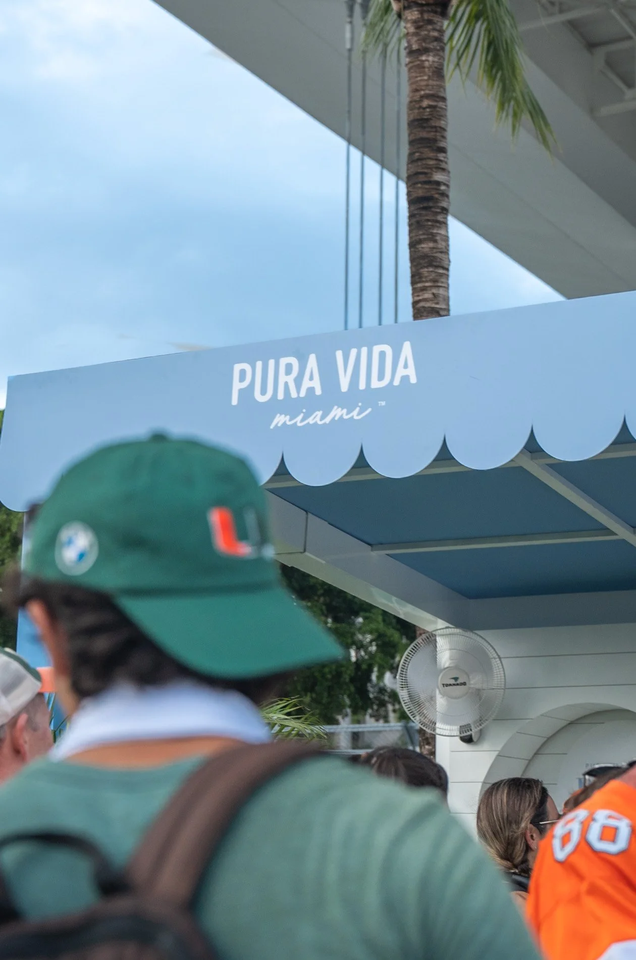 People waiting in line outside a Miami location with a blue awning labeled "Pura Vida Miami" and a palm tree in the background.