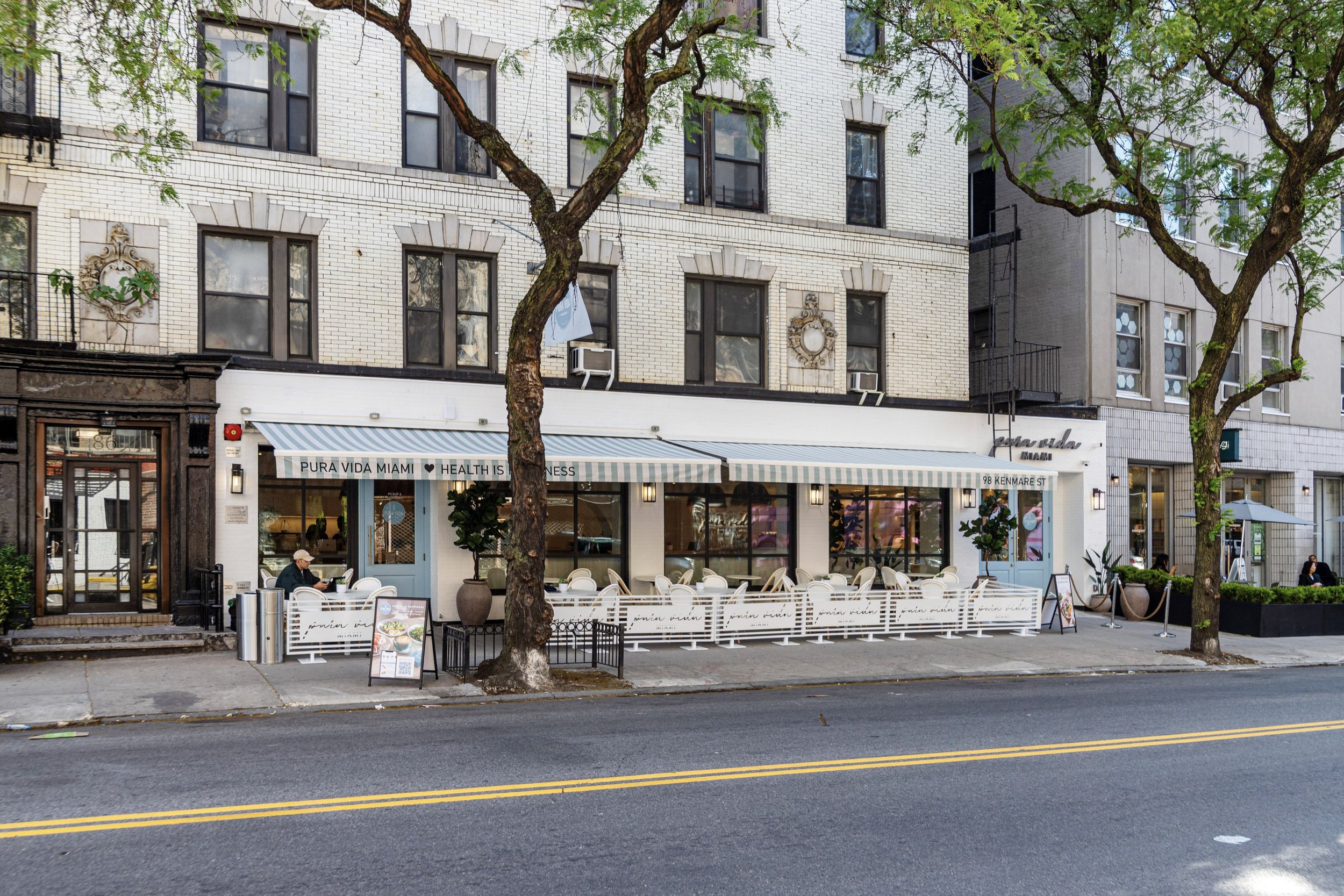 Exterior of a restaurant with outdoor seating on a city sidewalk, featuring white chairs, potted plants, and a striped awning, with tall trees and multi-story buildings in the background.