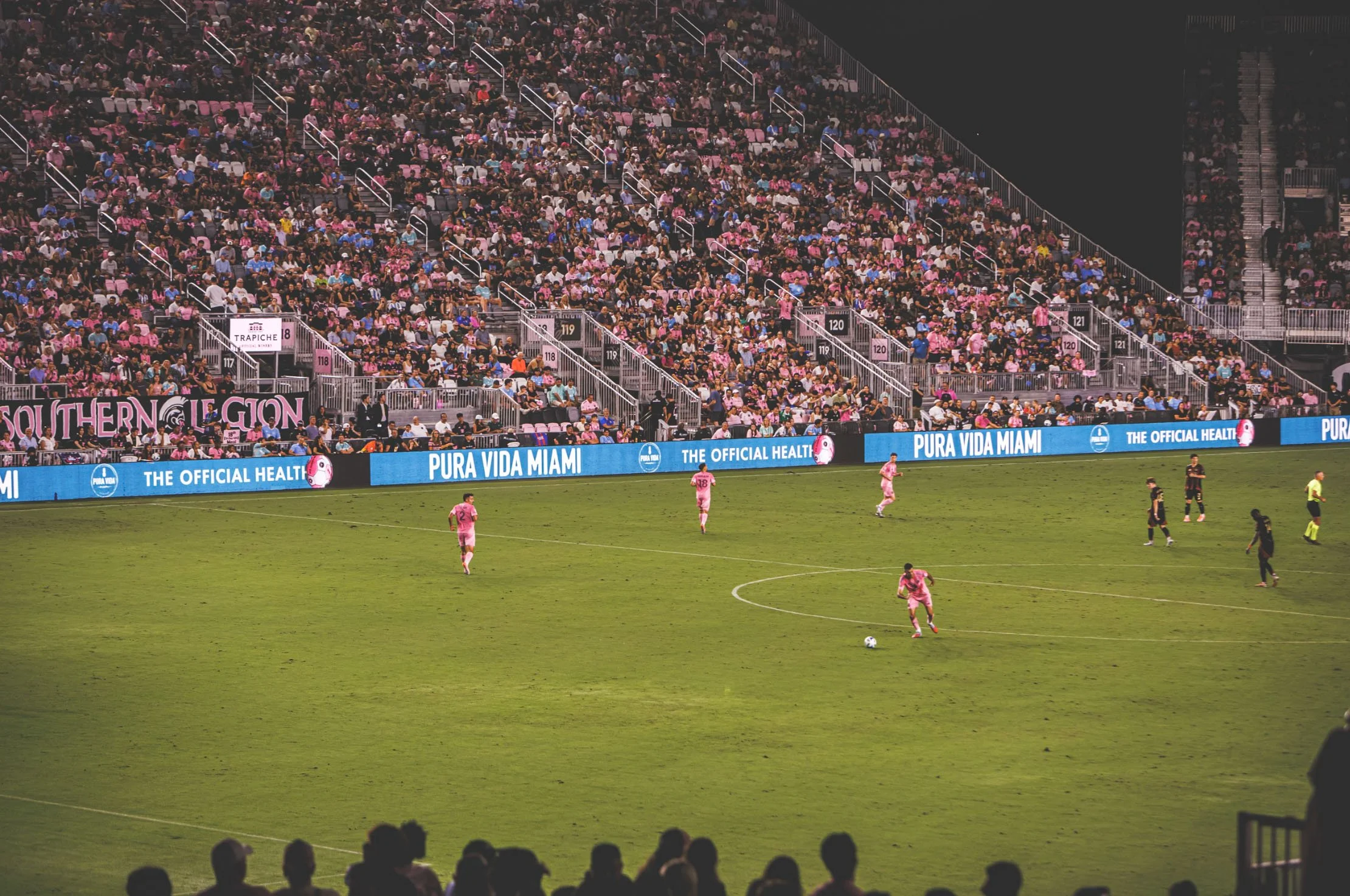 A soccer match in a stadium filled with a large crowd of spectators. Several players are on the field, some in pink uniforms and others in black, with an ad board reading 'Pura Vida Miami' visible along the sideline.