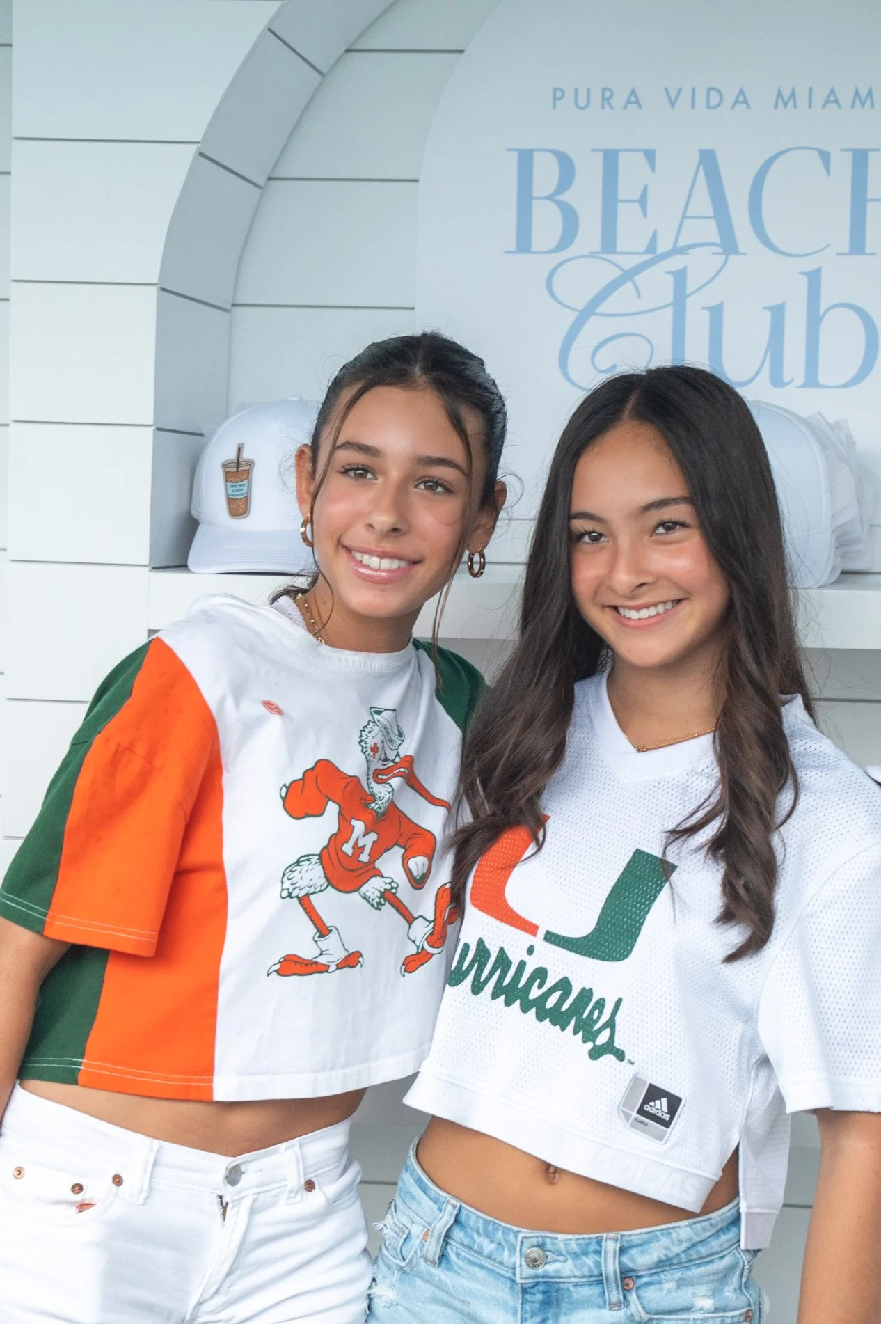 Two teenage girls smiling at a Miami Beach Club event, wearing Miami Hurricanes football jerseys, with Miami Beach Club signage in the background.