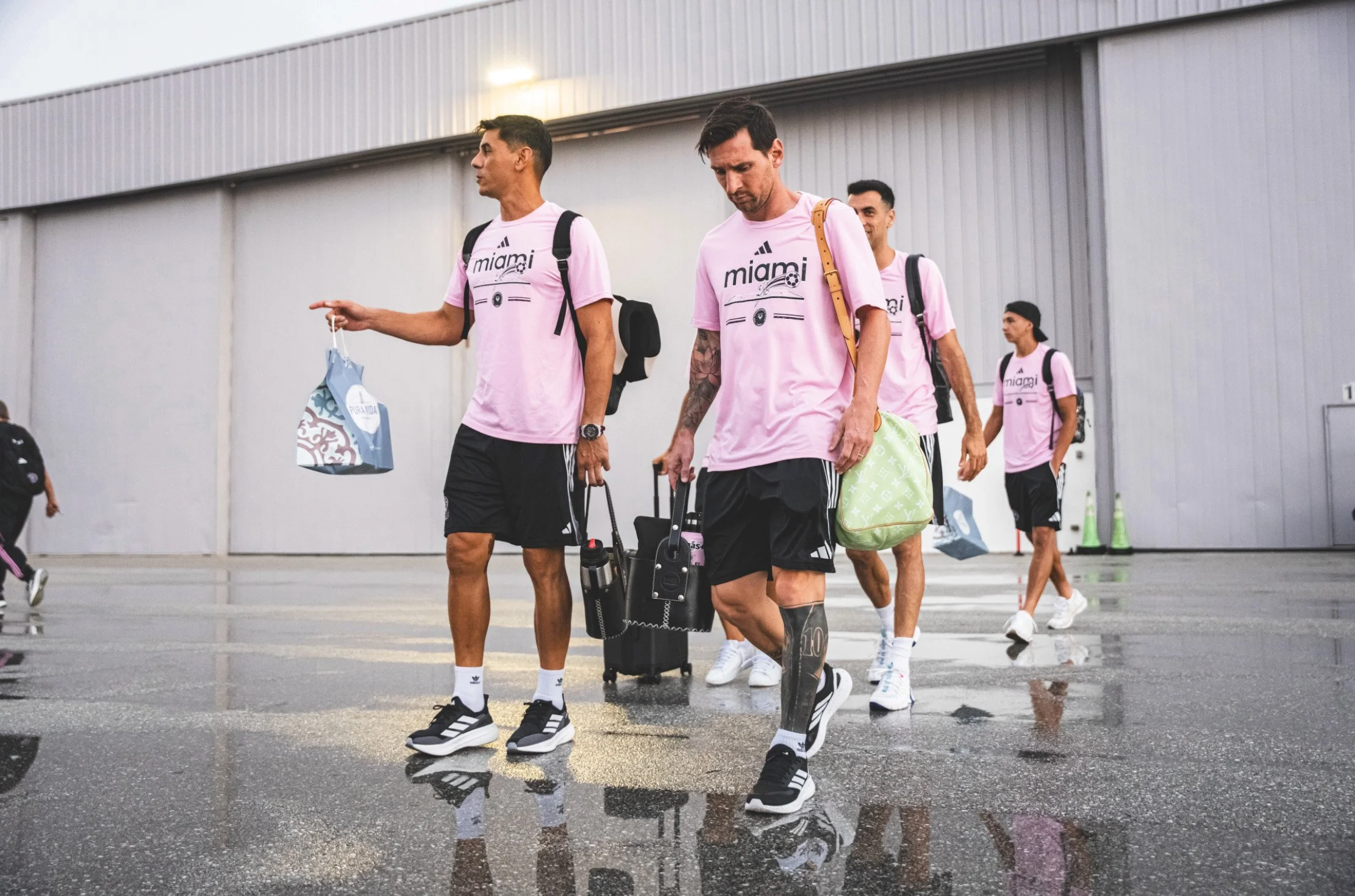 A group of five men walking outside an airport wearing matching pink Miami soccer jerseys, carrying luggage and bags.