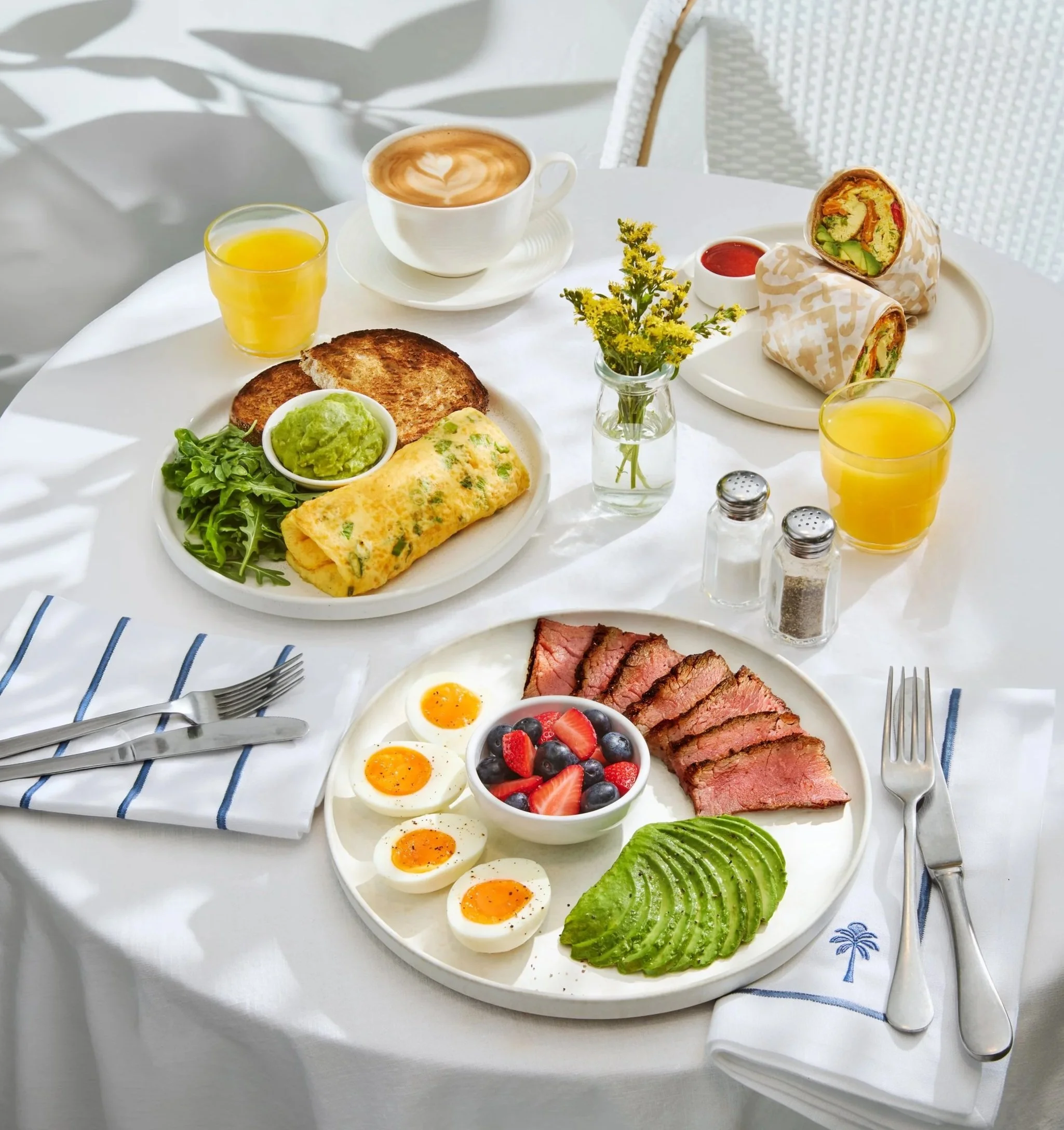 Breakfast table spread including Scallion & Feta Omelette, Steak & Eggs. Breakfast Burrito, a cappuccino and orange juice.